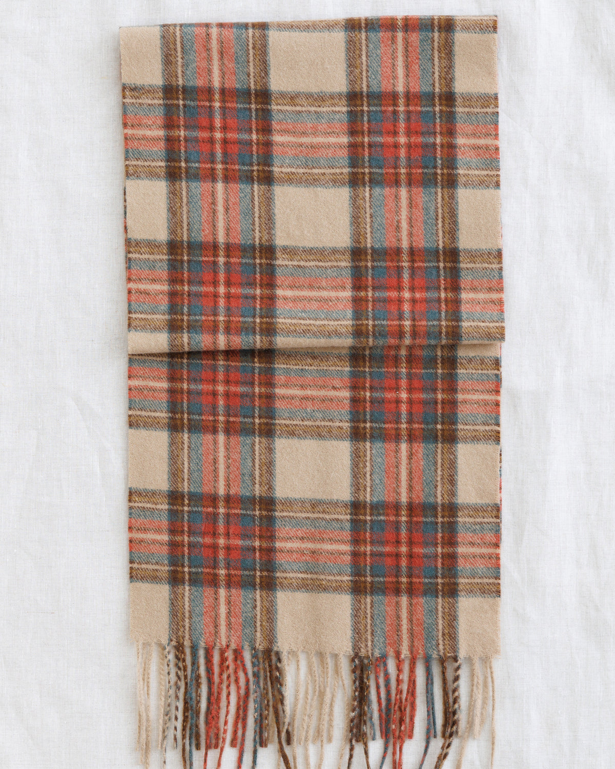 A folded plaid scarf in beige, rust red, brown, and muted blue-gray, with fringed ends, laid flat on a white textured fabric background.