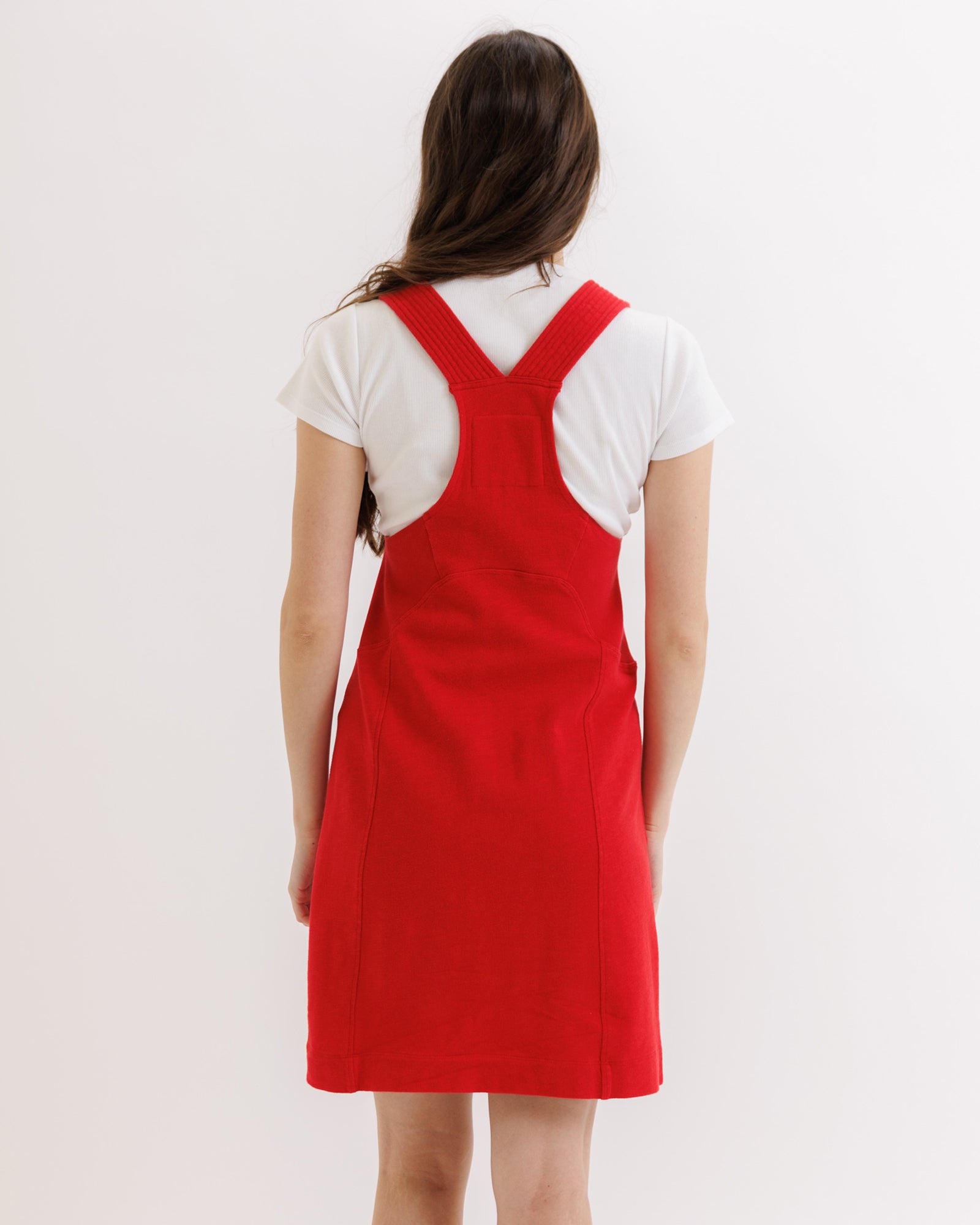 Person wearing a red pinafore dress over a white shirt on a plain background