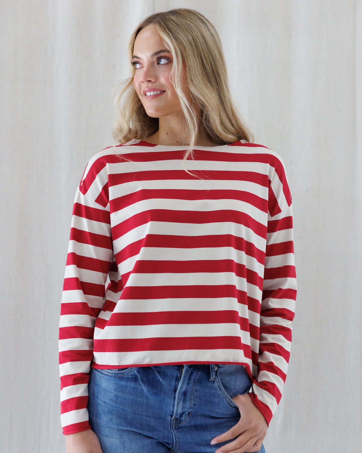 A woman wearing a red and off-white horizontally striped long-sleeve top with a wide boat neckline, paired with blue jeans, standing against a light neutral curtain background.
