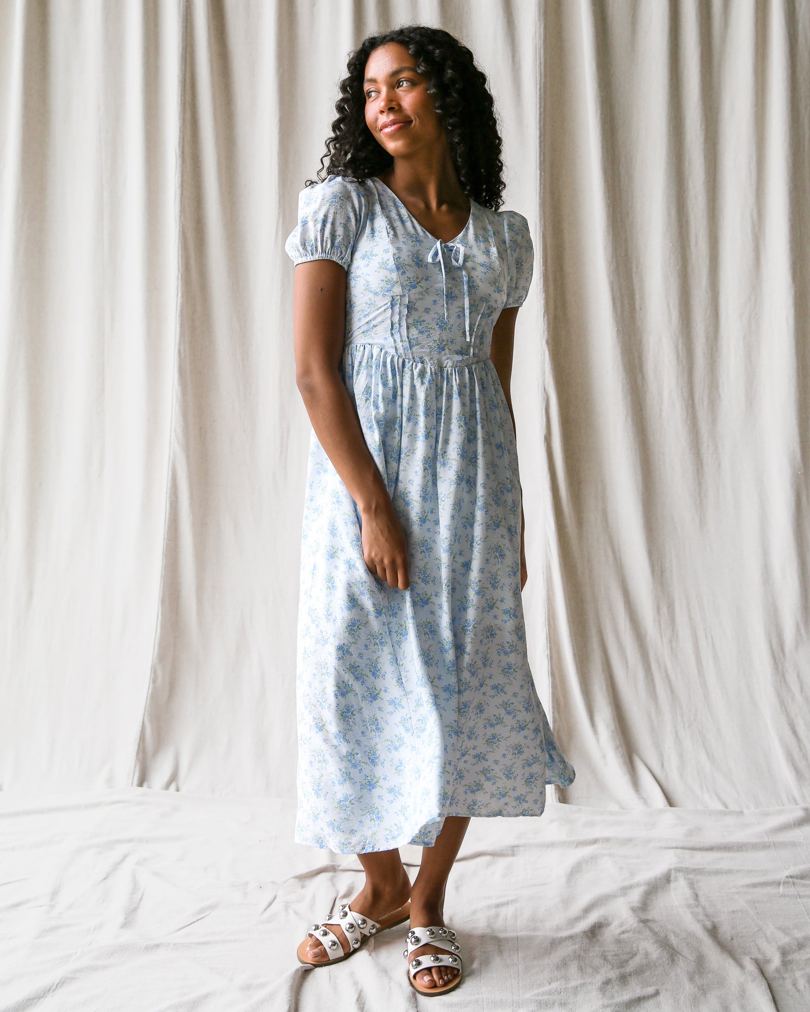 Woman wearing a light blue floral dress standing against a white curtain.