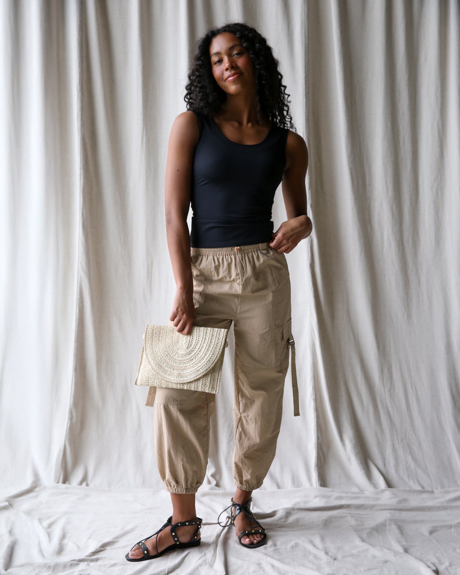 Woman wearing a black tank top and beige cargo pants, holding a beige clutch against a white curtain background.