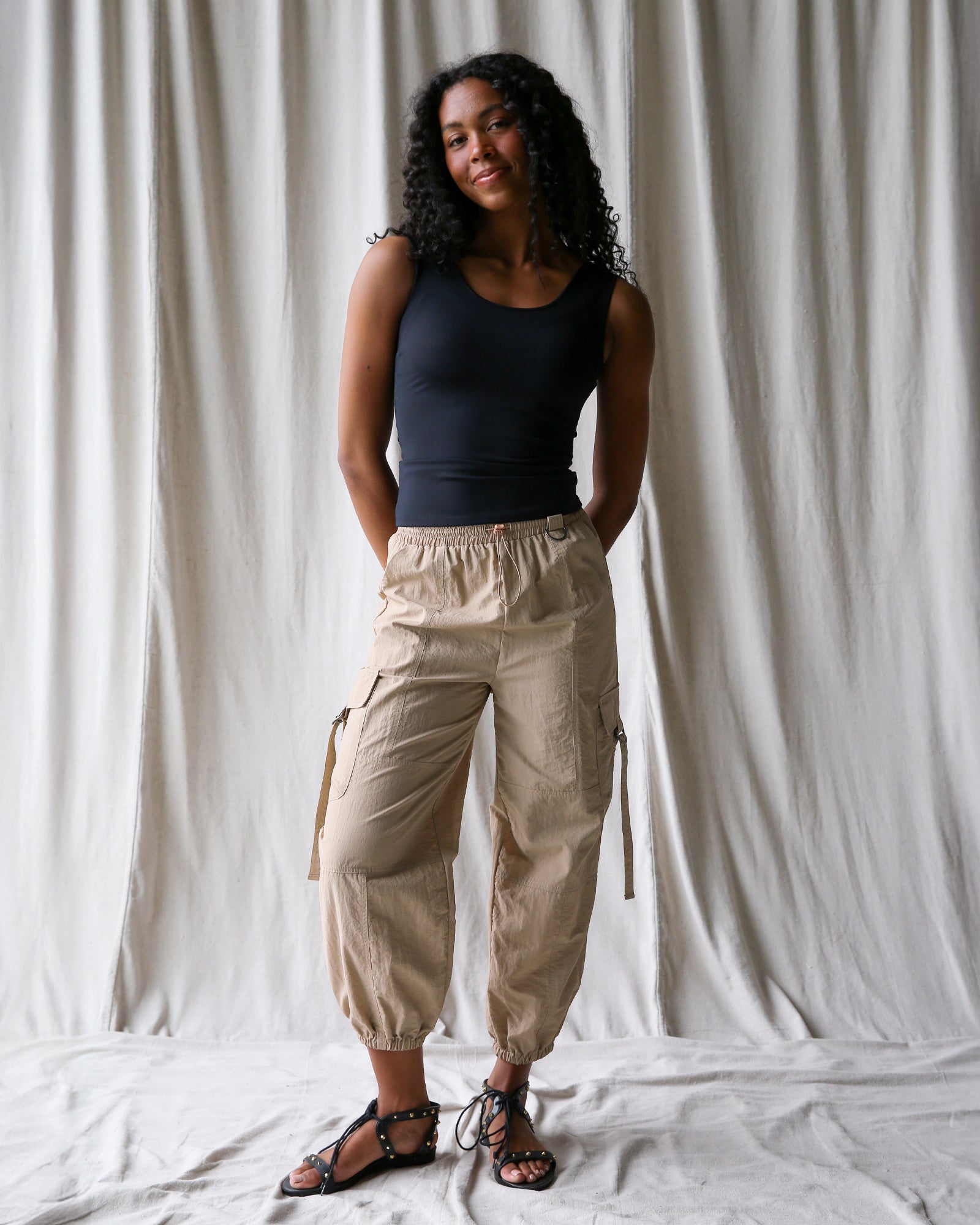 Woman wearing a black tank top and beige cargo pants standing against a white curtain.