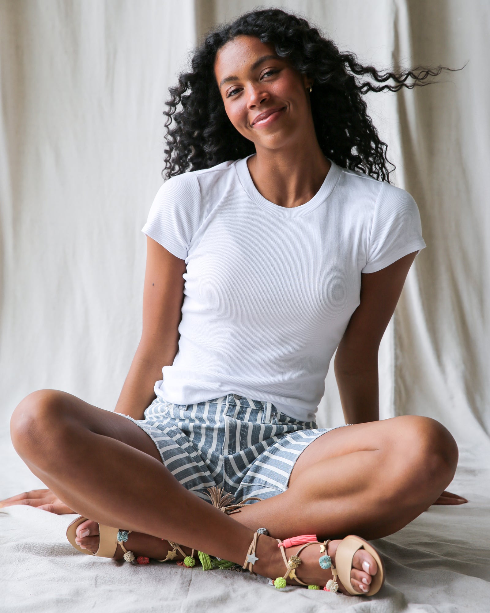 Woman sitting cross-legged on a bed wearing a white t-shirt and striped shorts.