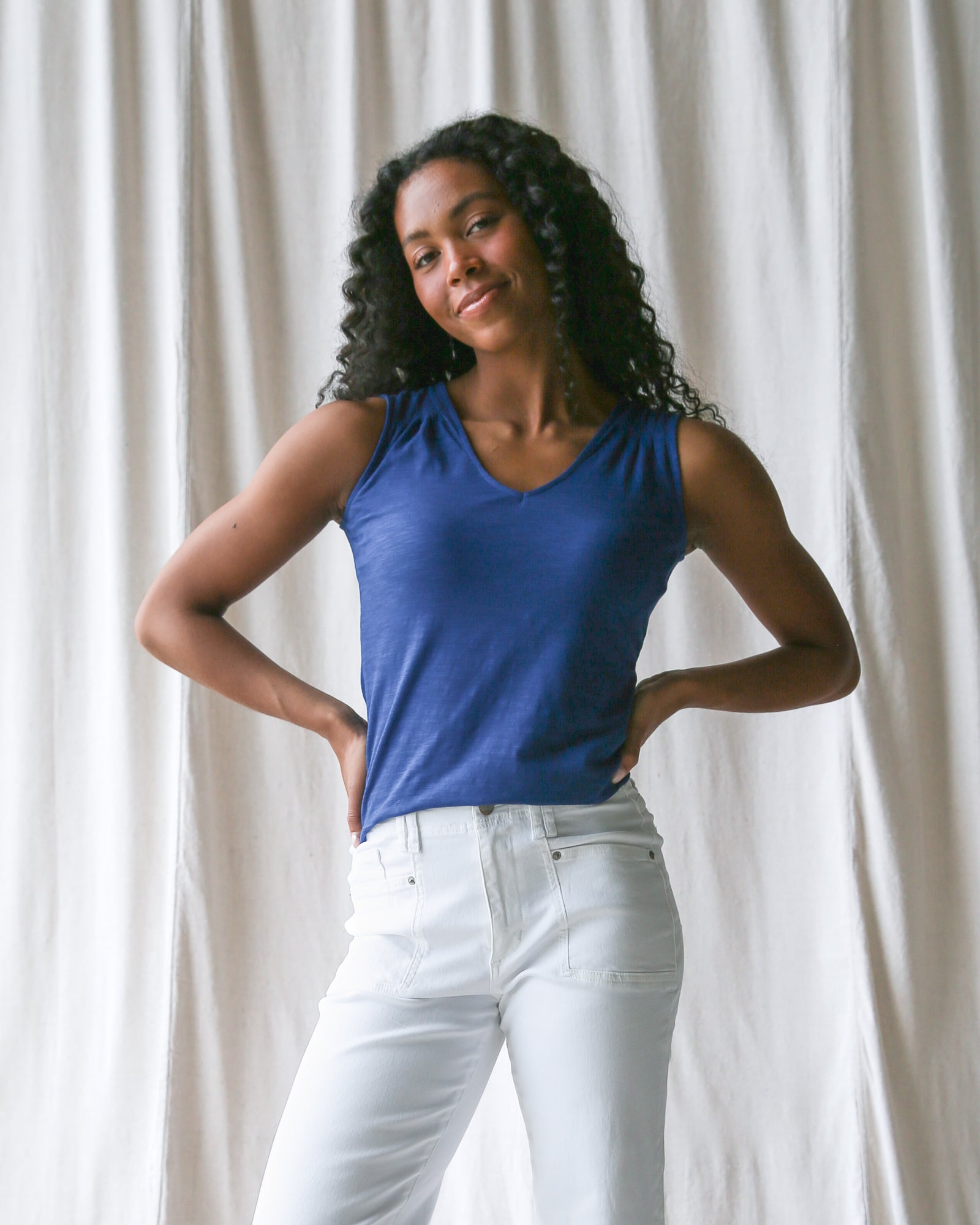 Woman wearing a blue tank top and white pants against a white curtain background