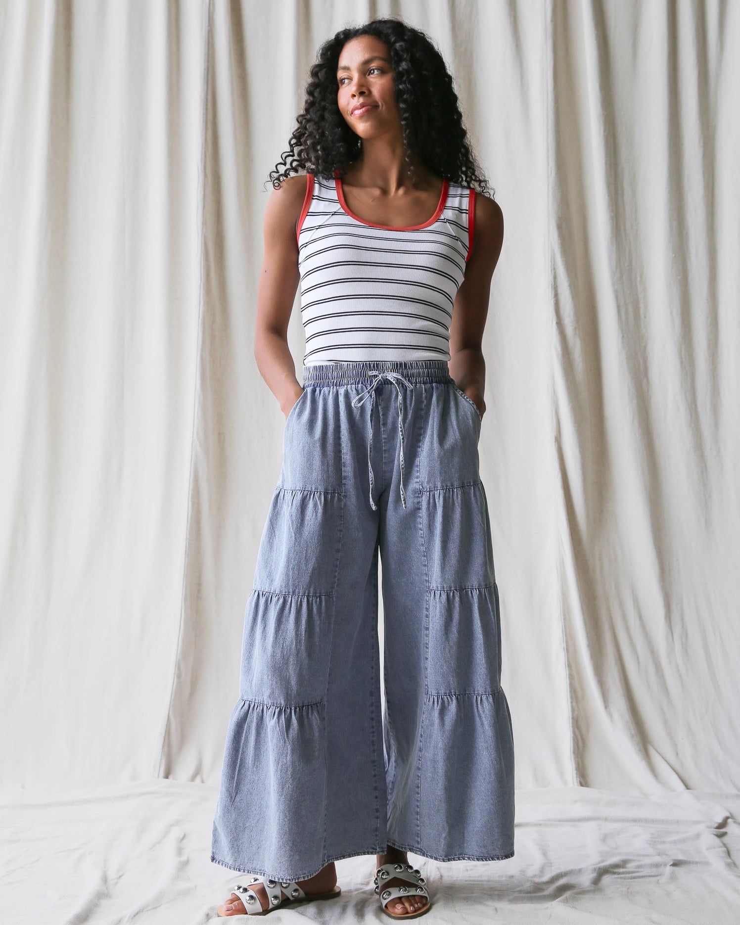 Woman wearing a striped tank top and blue tiered pants against a white curtain background