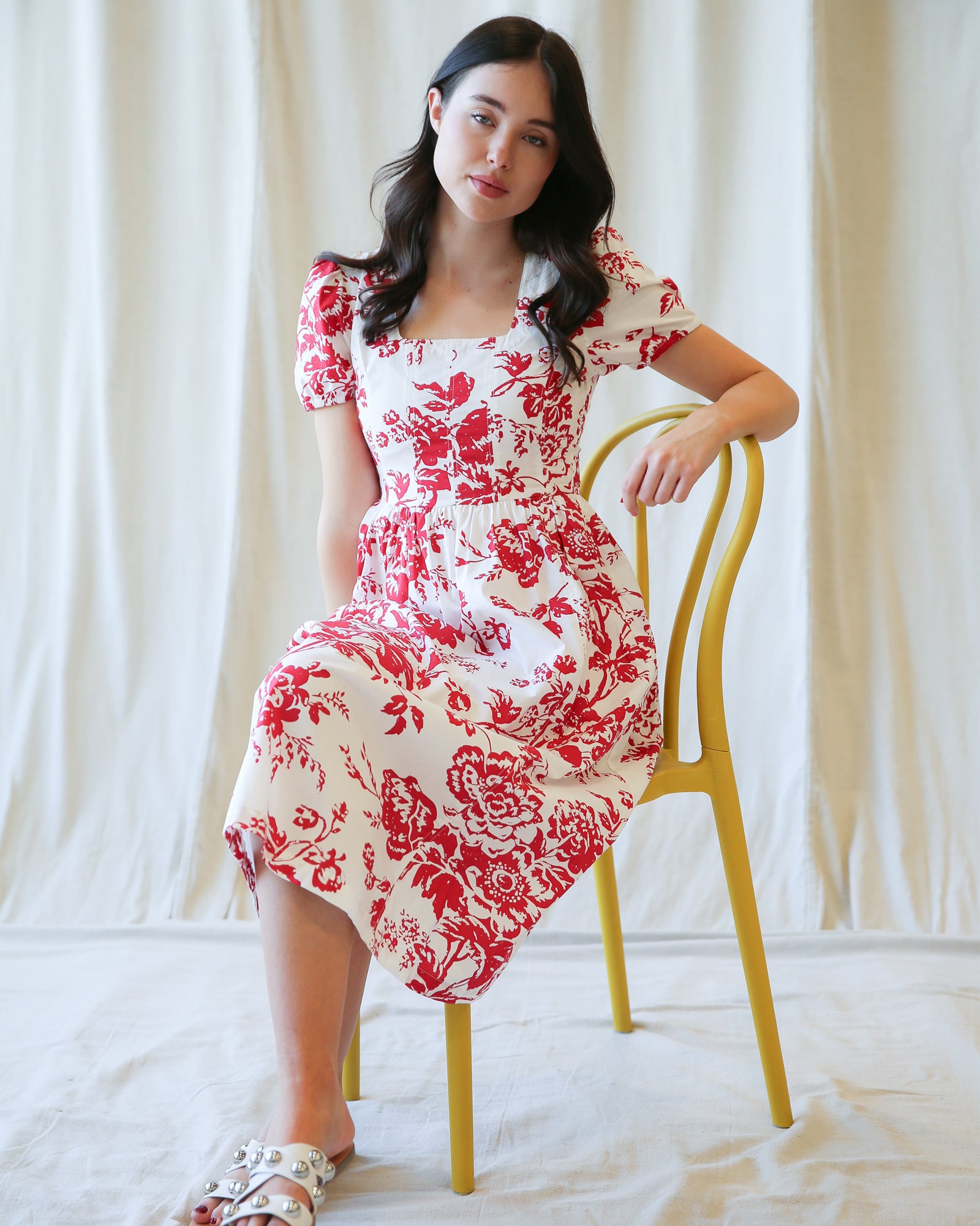 Woman in a red and white floral dress sitting on a yellow chair against a white curtain background