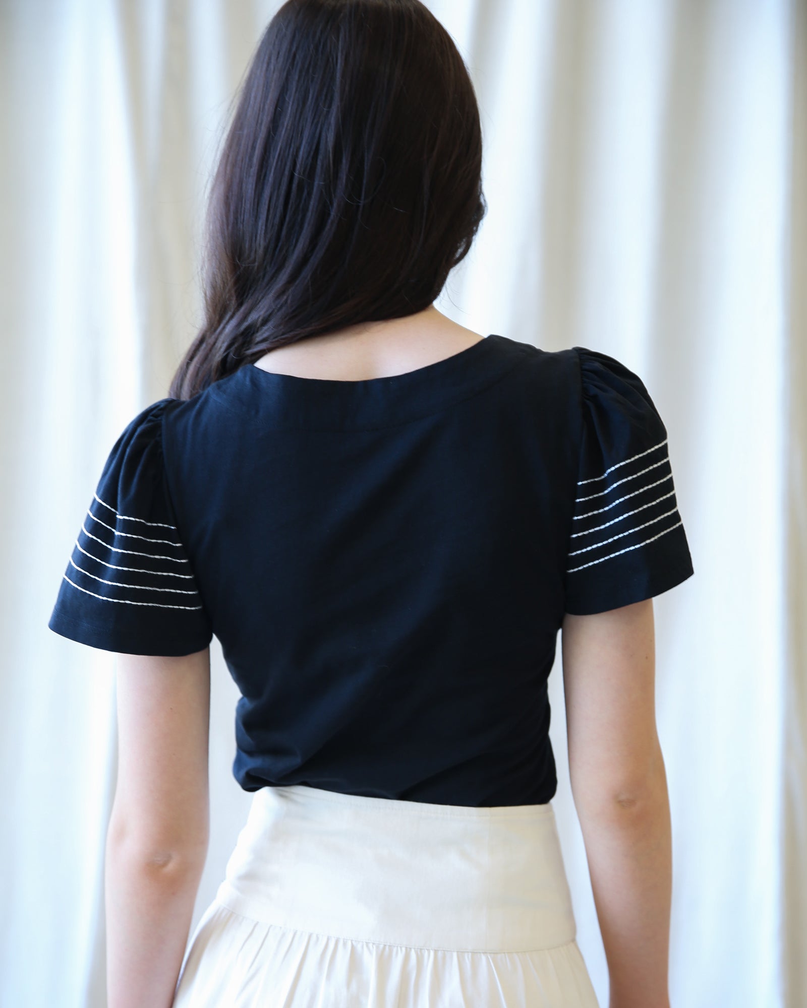 Person wearing a black top with white stripes on sleeves against a white curtain background