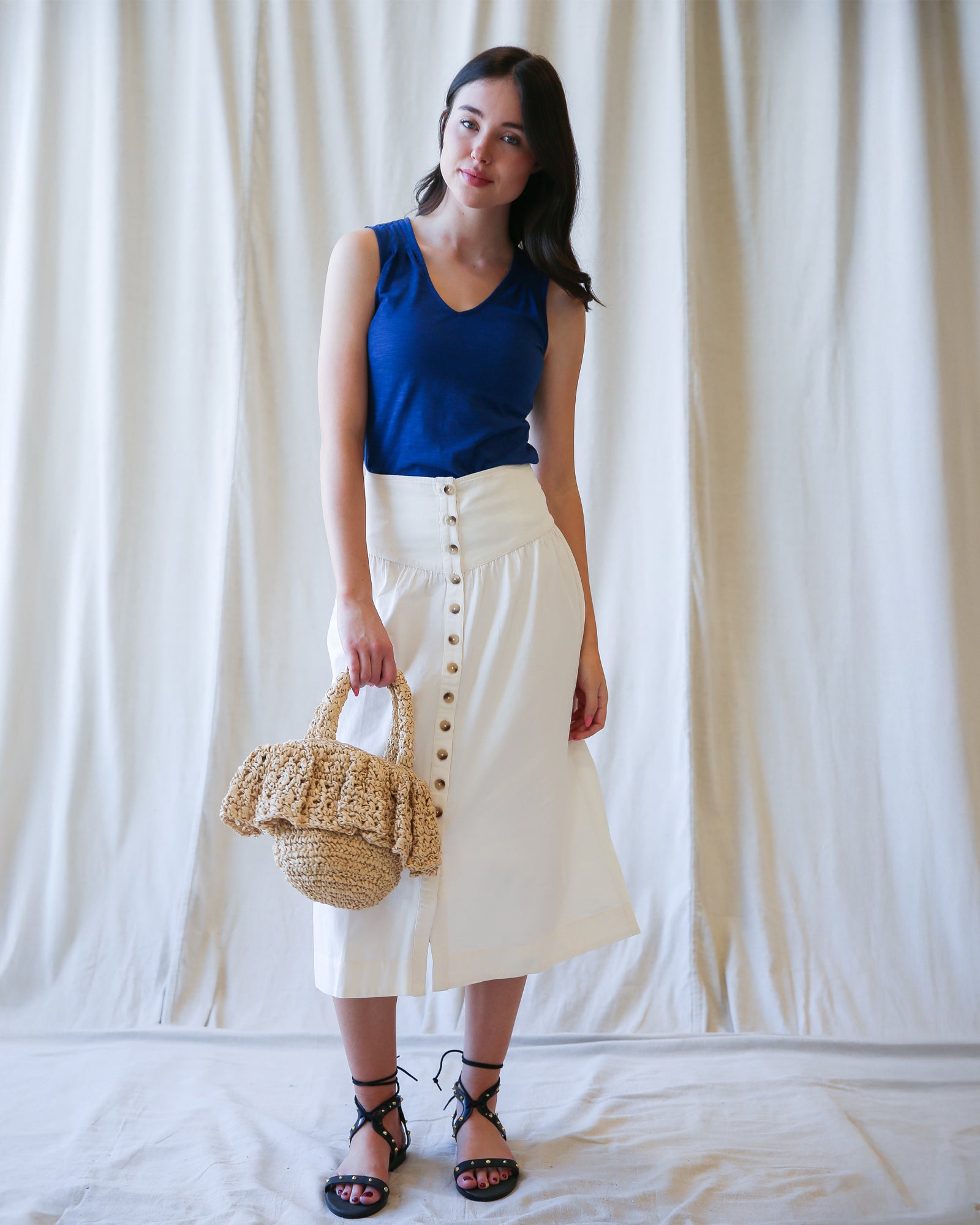Woman wearing a blue top and white skirt holding a straw bag against a white curtain background