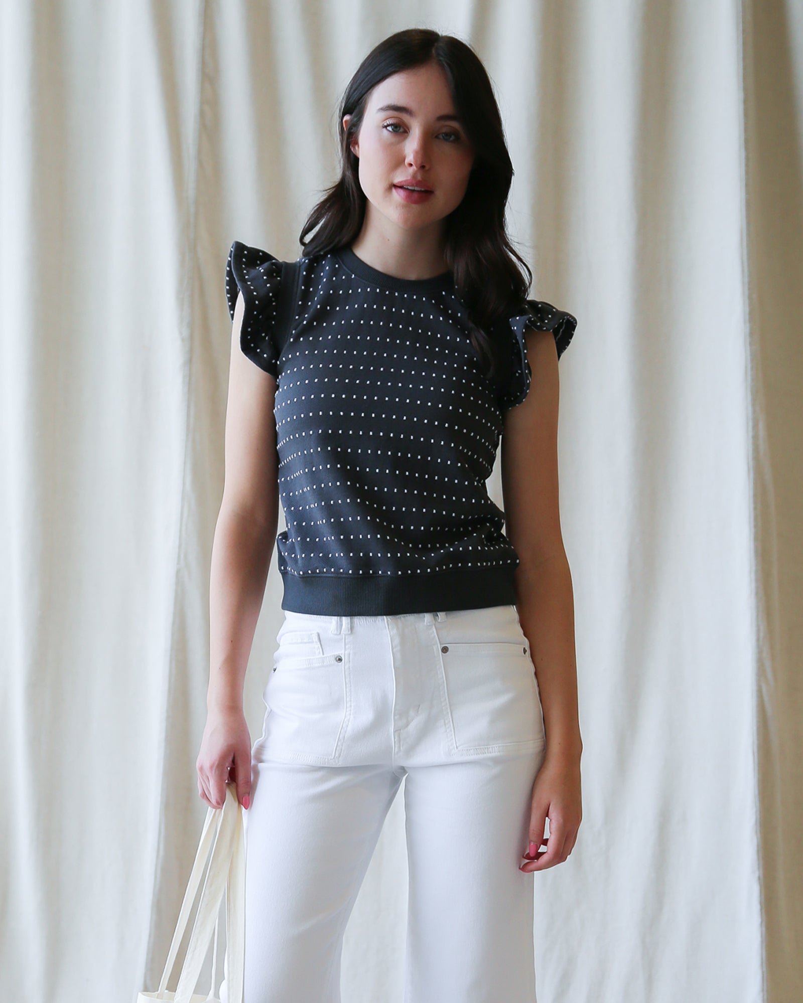 Woman wearing a black top with white polka dots and white pants against a white curtain background