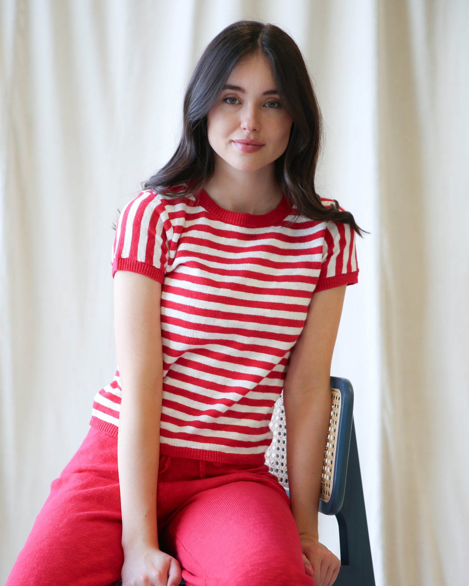 Woman wearing a red and white striped shirt and pink pants sitting against a white curtain.