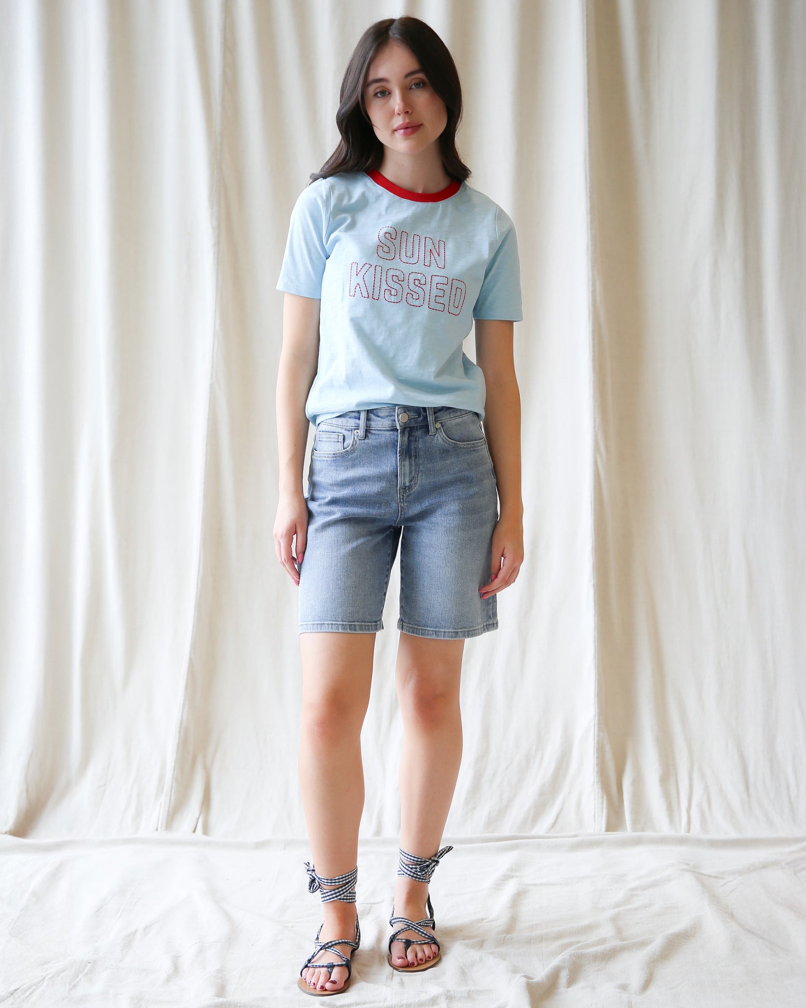 Woman wearing a light blue t-shirt with 'Sun Kissed' text and denim shorts against a white background