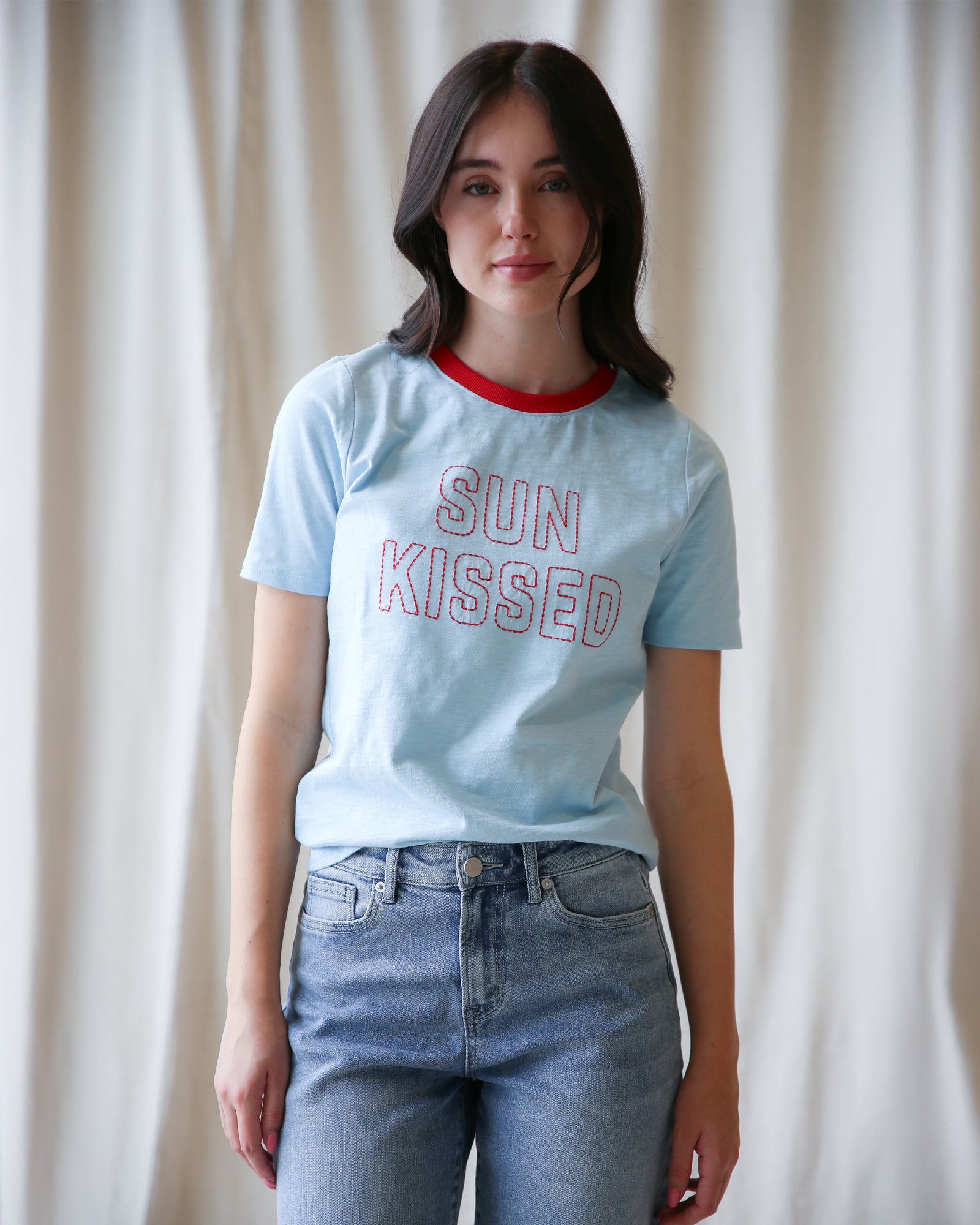 Person wearing a light blue t-shirt with 'SUN KISSED' text and jeans against a white curtain background