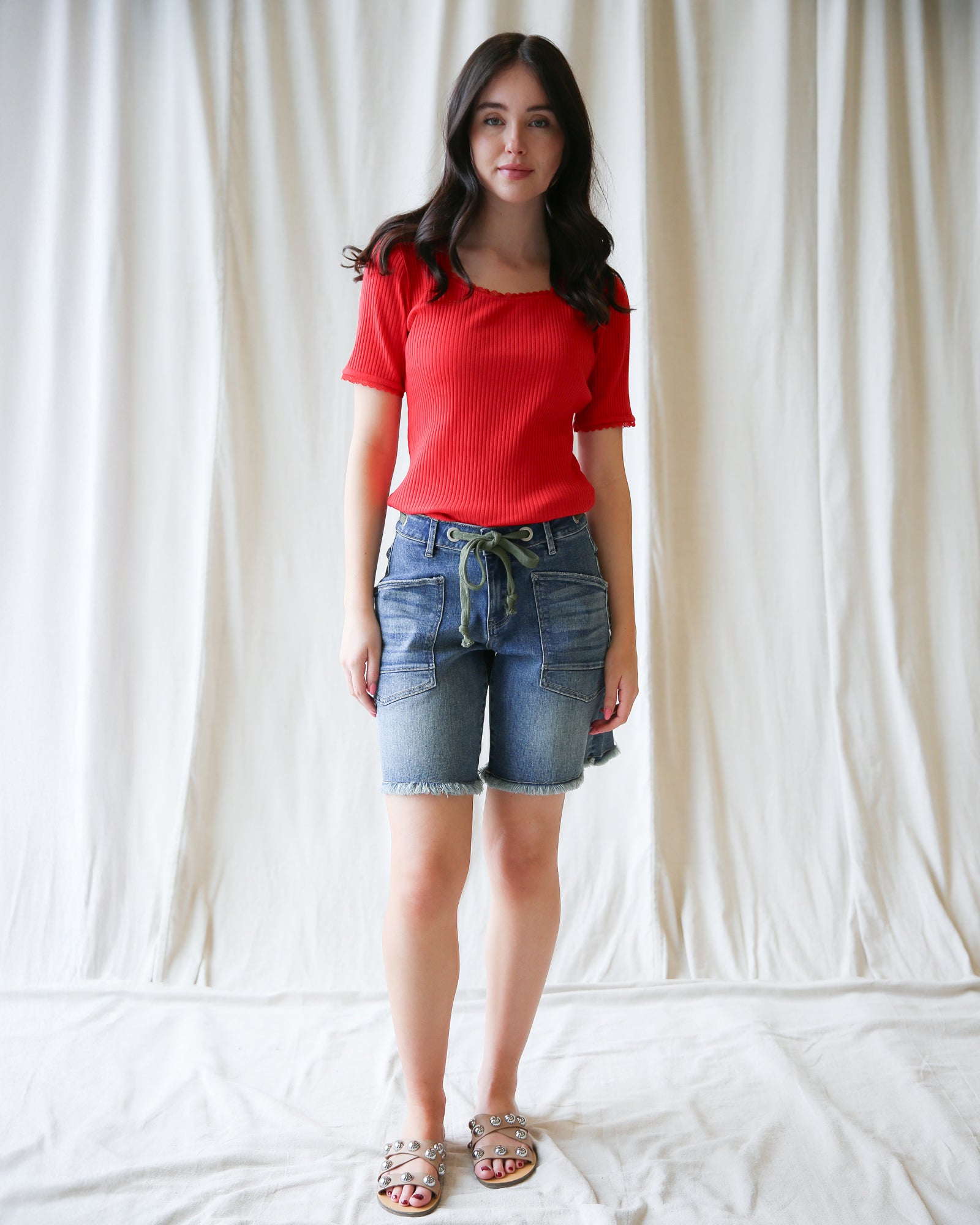 Woman wearing a red top and denim shorts against a white curtain background