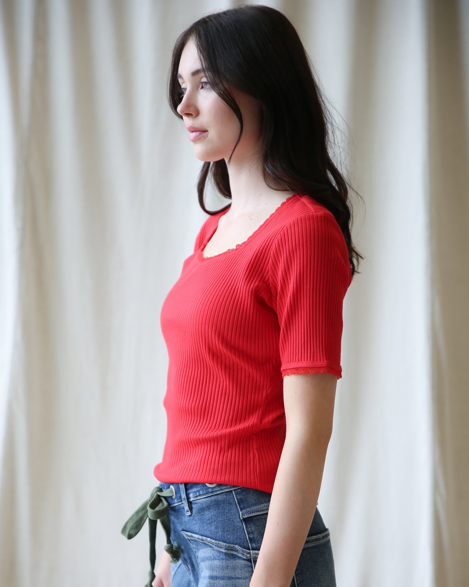 Woman wearing a red top and blue jeans against a white curtain background