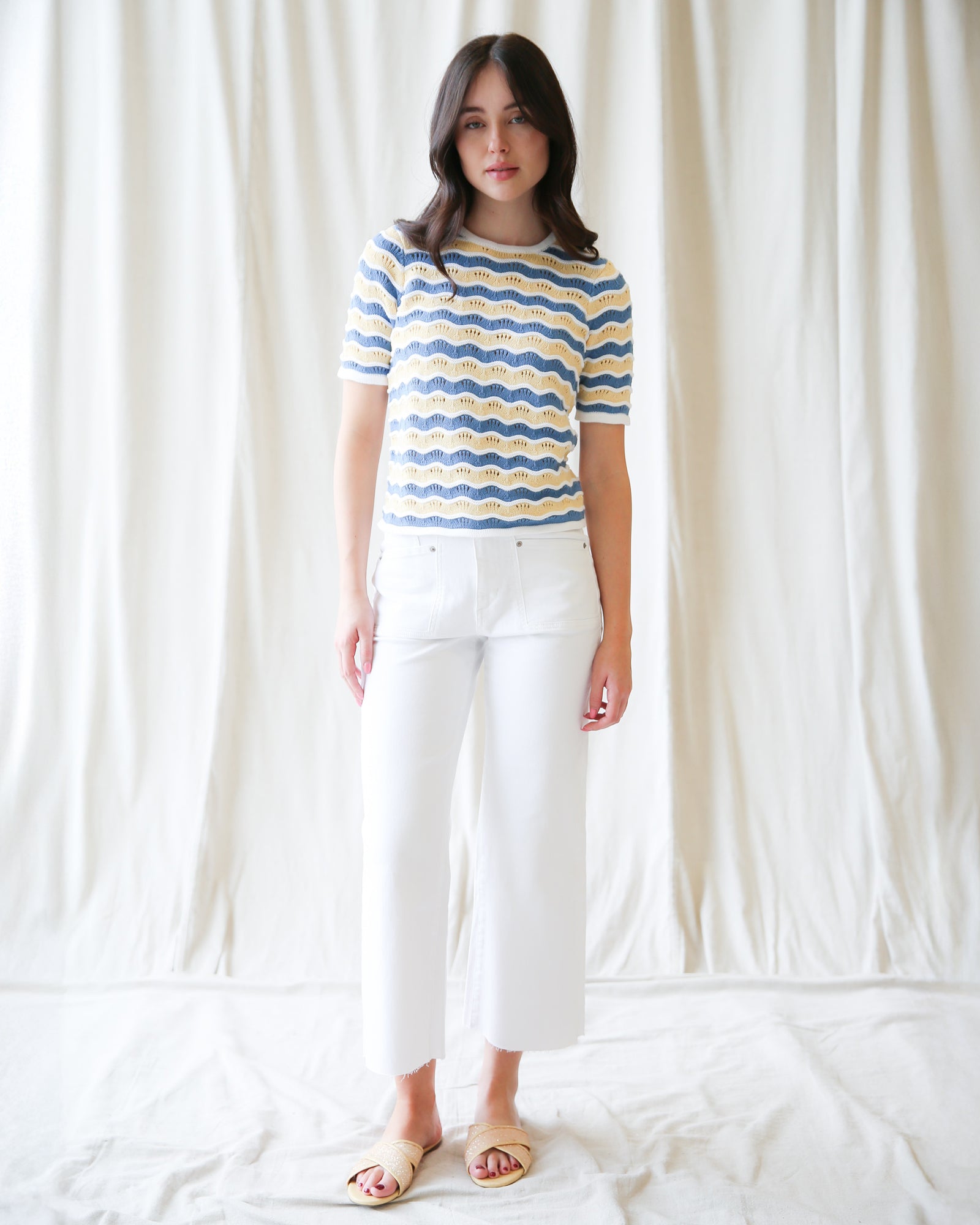 Woman wearing a blue and yellow striped shirt and white pants against a white curtain background