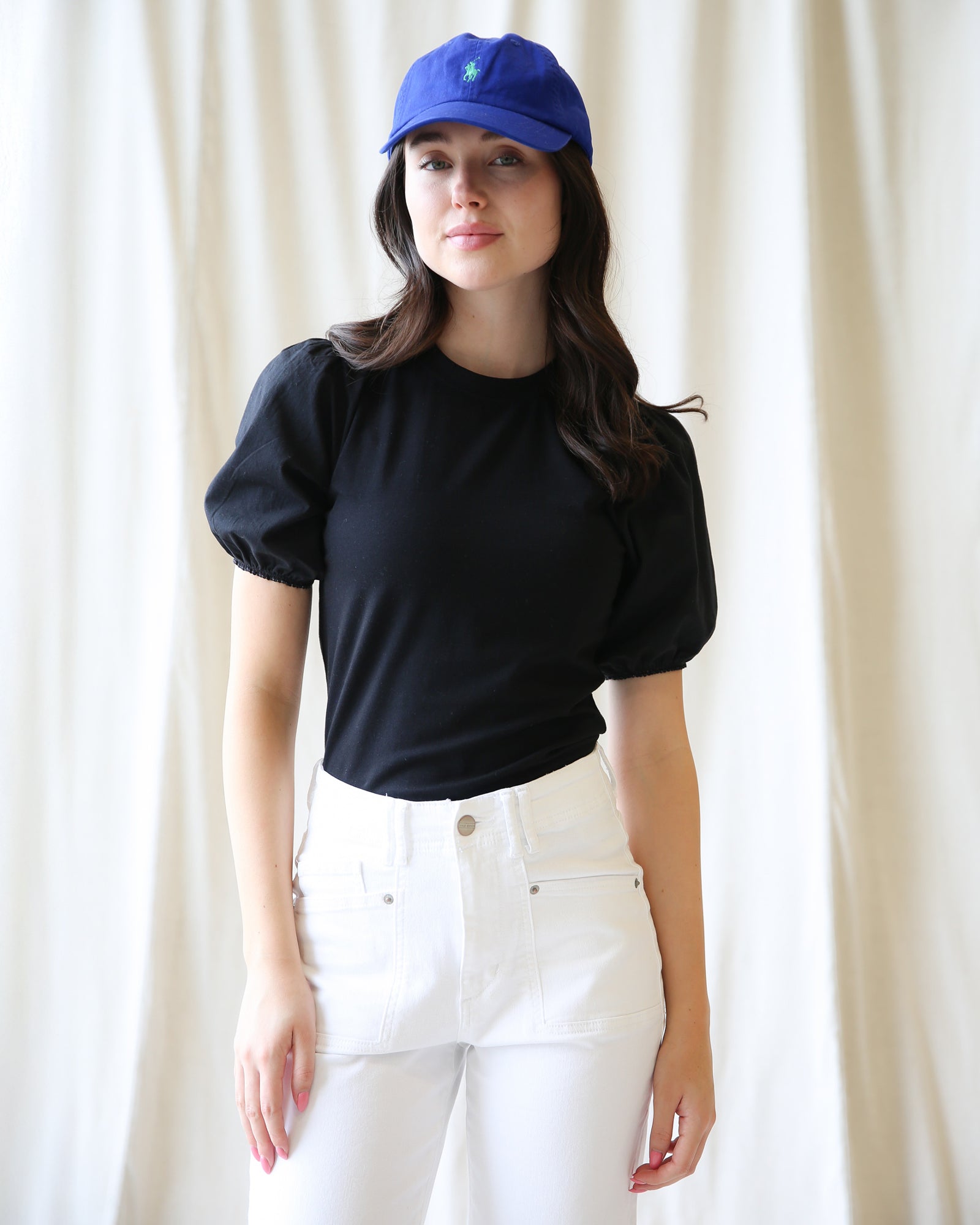 Woman wearing a black top and white pants with a blue cap against a white curtain background