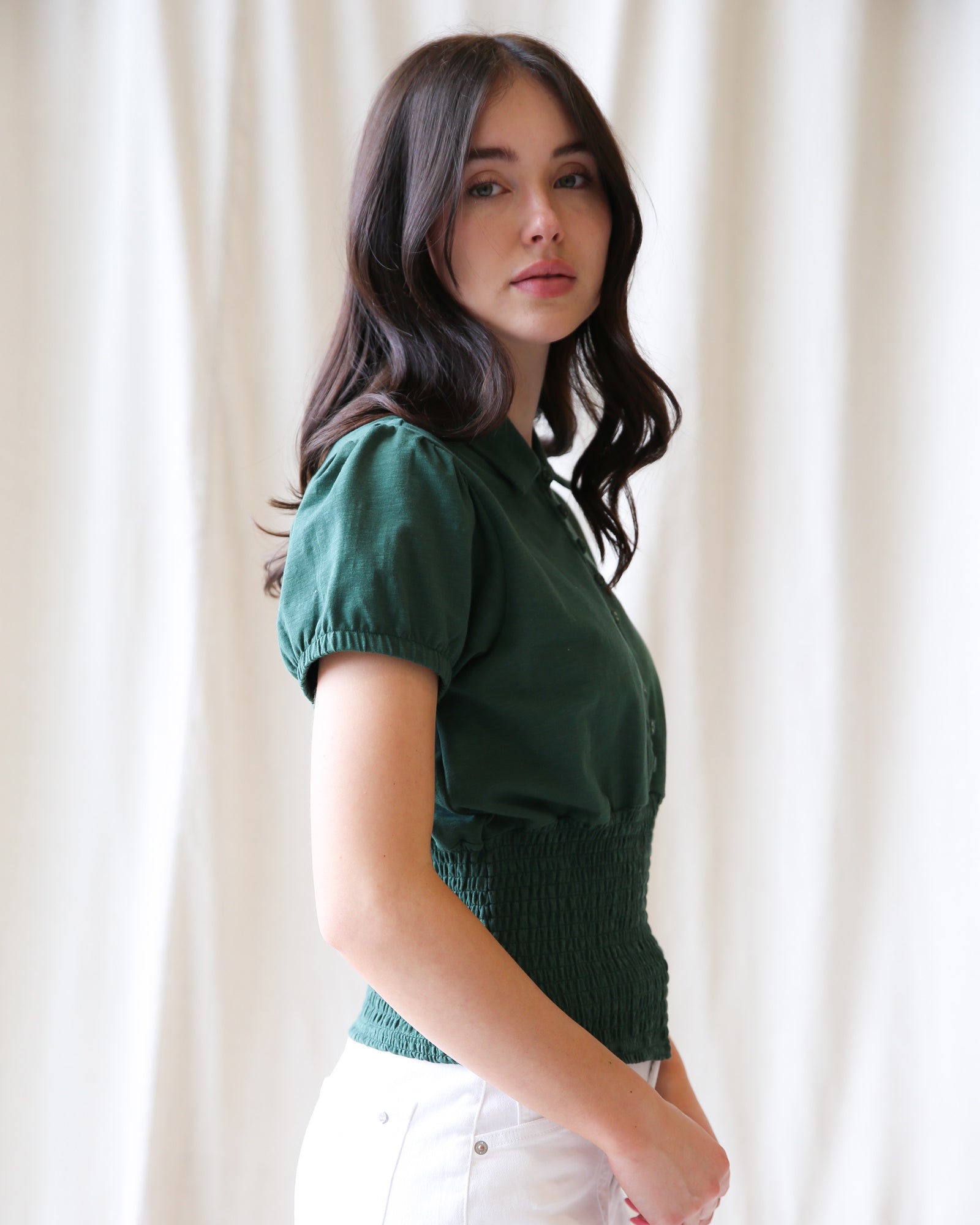 Woman wearing a green top and white pants against a light curtain background