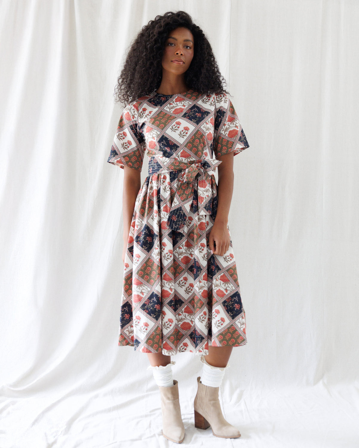 A woman modeling a short-sleeve midi dress with a multicolor patchwork diamond pattern in beige, navy, red, and white, styled with white socks and beige ankle boots against a white draped fabric background.