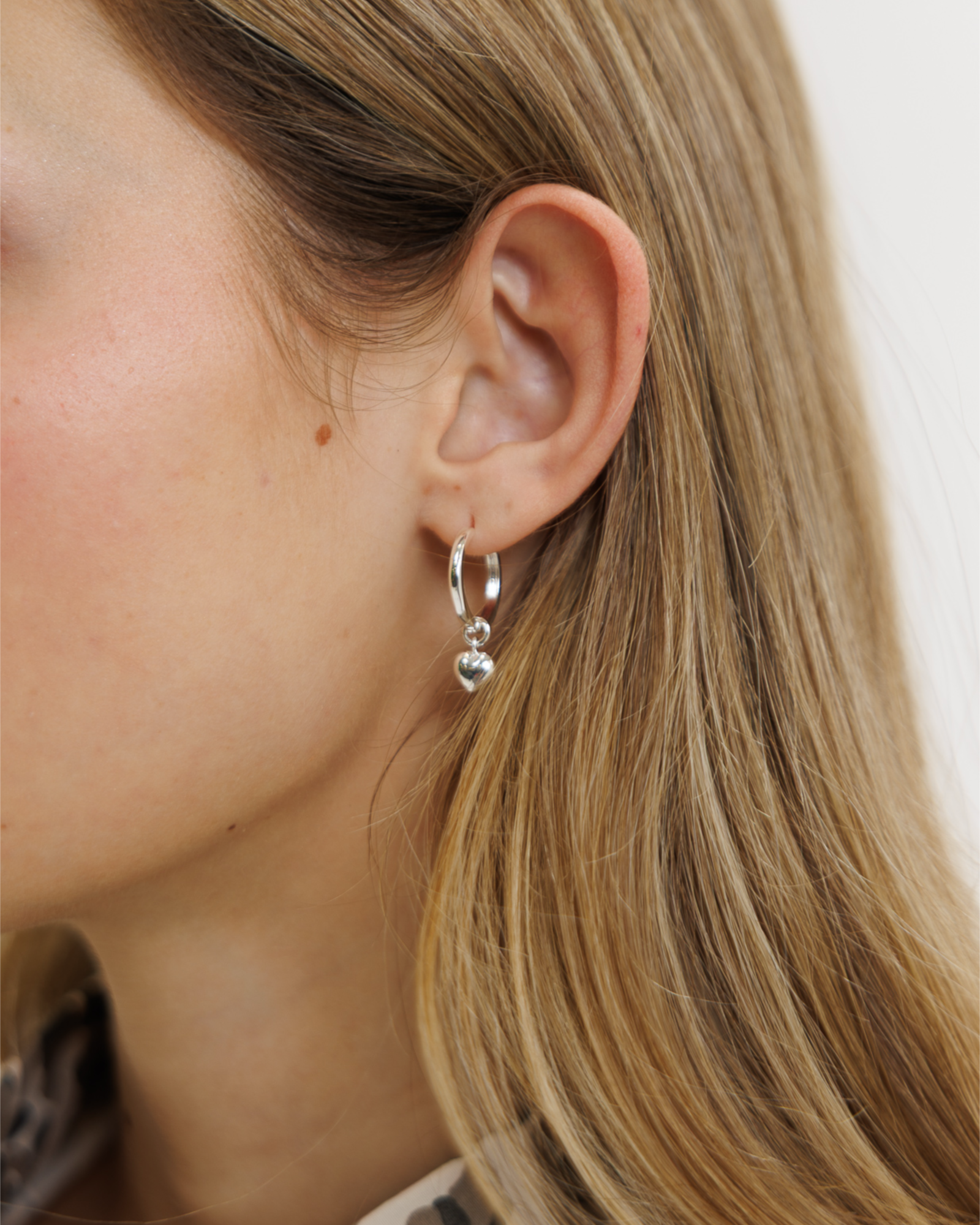 Close-up of a woman wearing silver hoop earrings with a heart charm on a neutral background