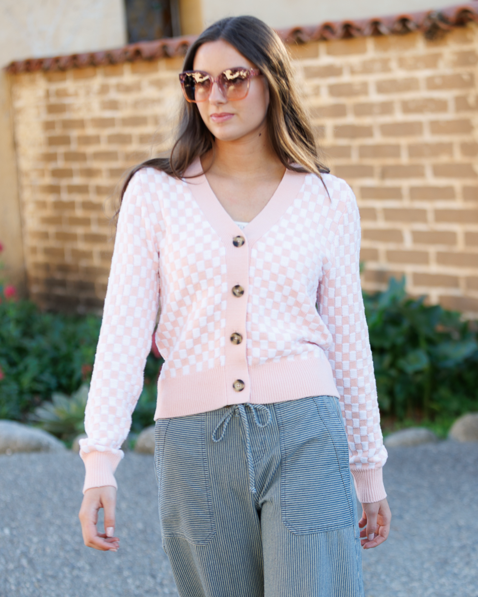 Woman wearing a pink checkered cardigan with buttons standing in front of a brick wall.