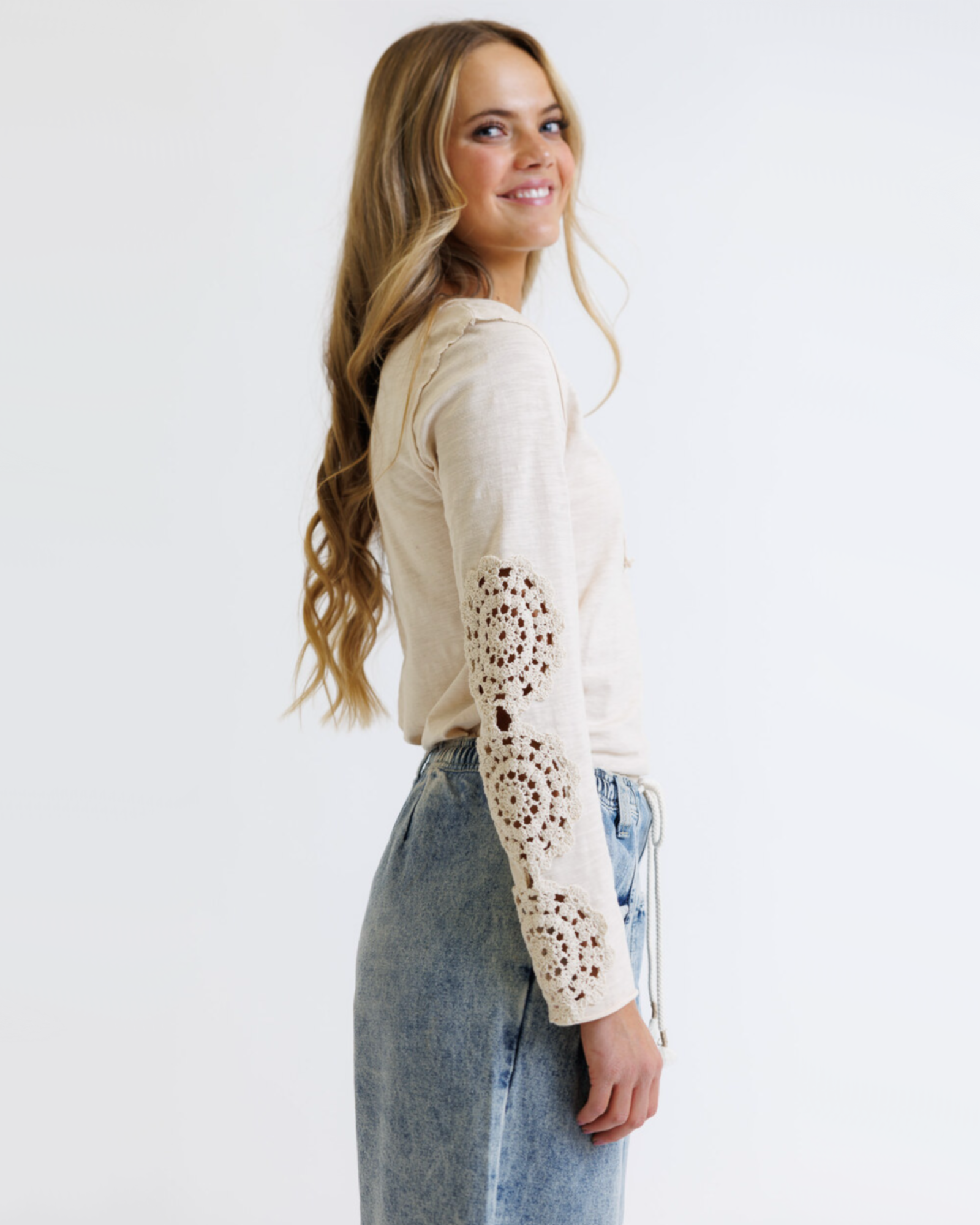 Woman wearing a cream colored long sleeve with crochet detailed sleeves against a white background