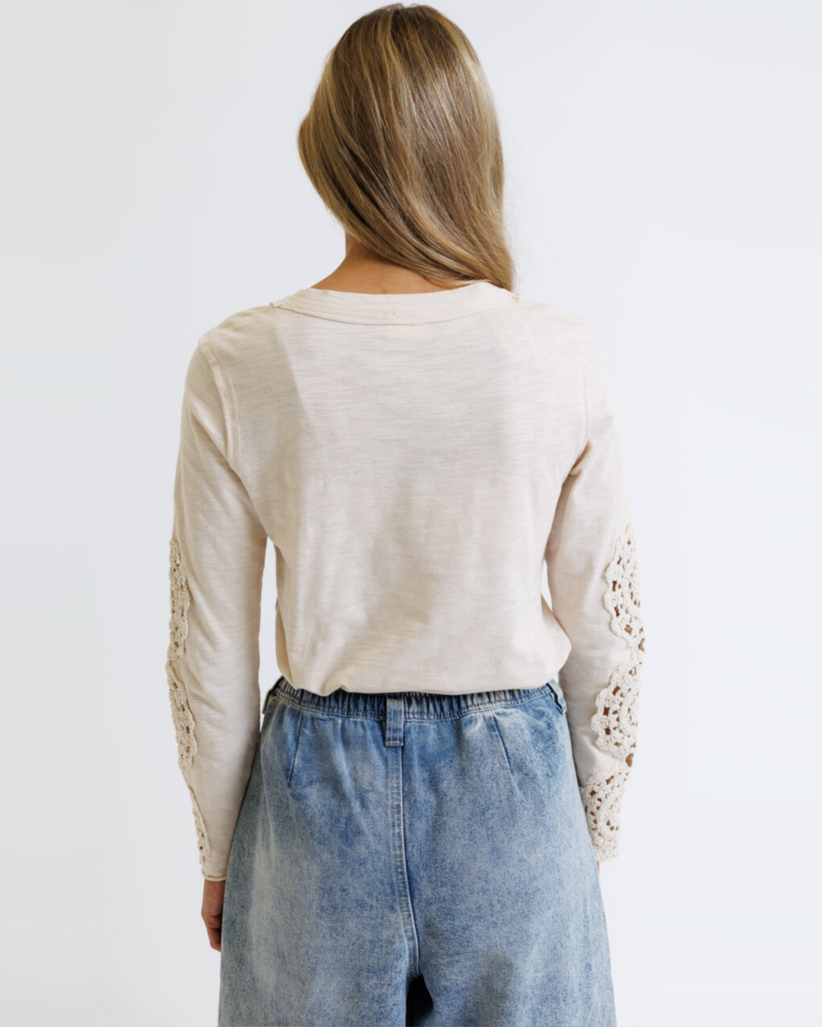 Woman wearing a cream colored long sleeve with crochet detailed sleeves against a white background