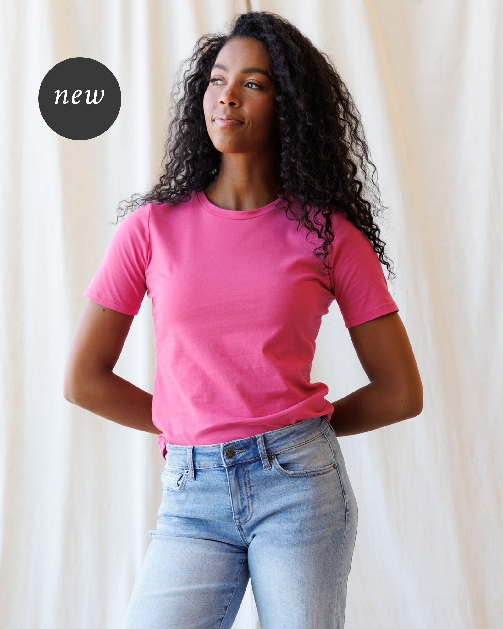 A woman wearing a pink short-sleeve crewneck t-shirt and light blue jeans, standing against a fabric backdrop.
