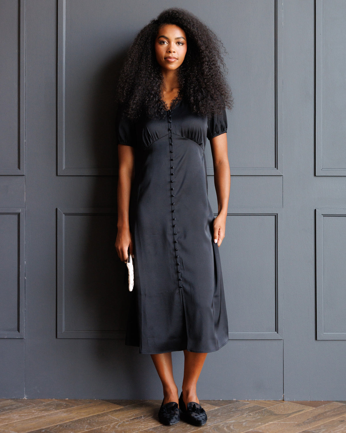 A woman with voluminous curly hair stands against a dark gray paneled wall wearing a dark charcoal gray midi dress with short sleeves, a V-neckline, and a central button placket. She is holding a small white clutch and wearing black pointed flats.