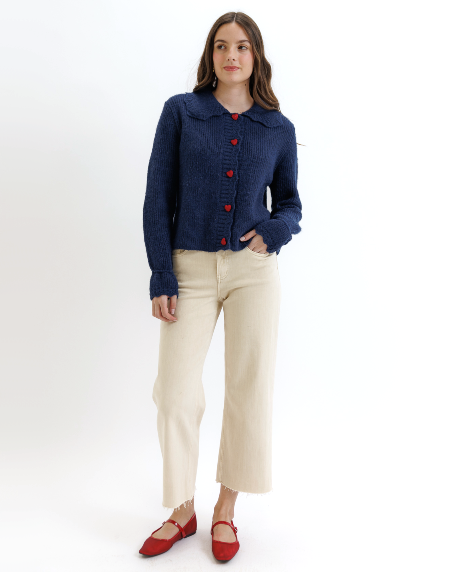 Woman wearing a navy blue cardigan with red buttons, beige pants, and red shoes on a white background