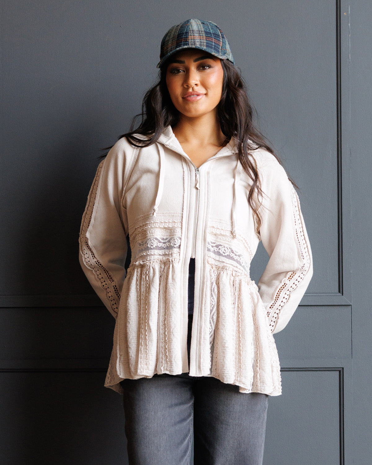 A woman wearing a cream-colored lace-trimmed zip-up hoodie with a peplum hem, paired with gray pants and a plaid cap, standing against a dark gray paneled wall.