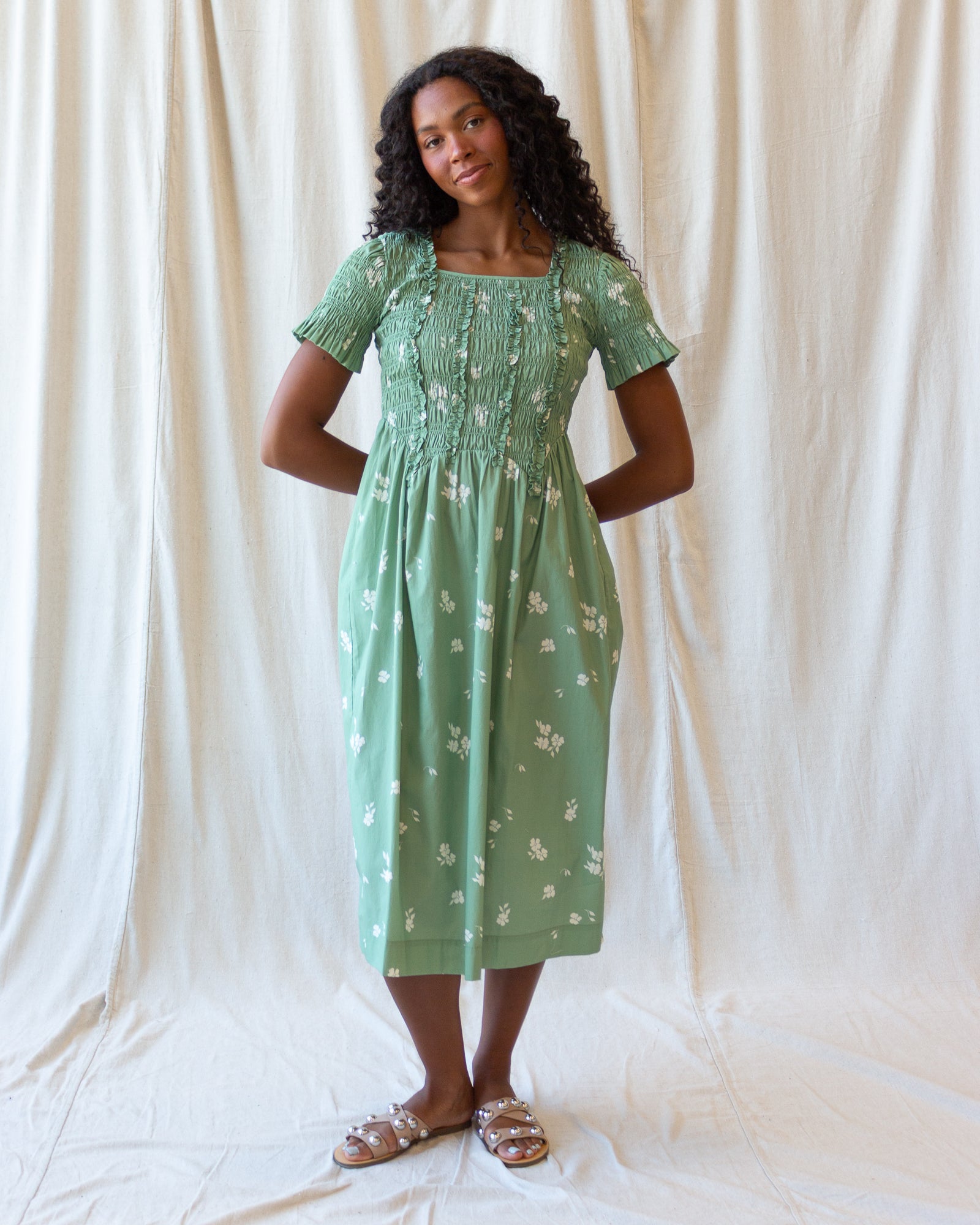 A woman modeling a light green midi dress with a white floral pattern, standing against a draped white fabric backdrop.