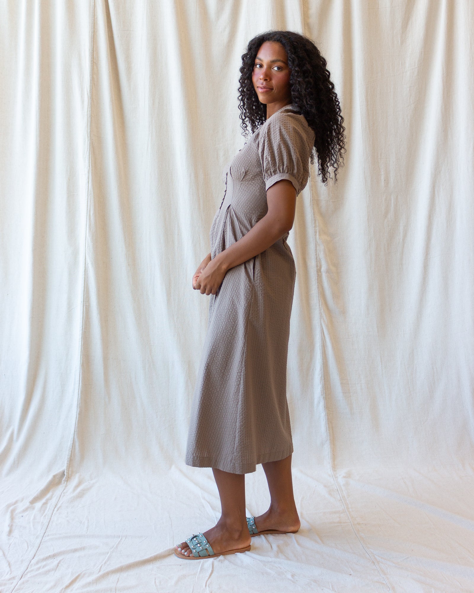 A woman stands in profile wearing a short-sleeve, knee-length taupe dress with puff sleeves and a gathered waist, posed against a draped cream fabric backdrop.