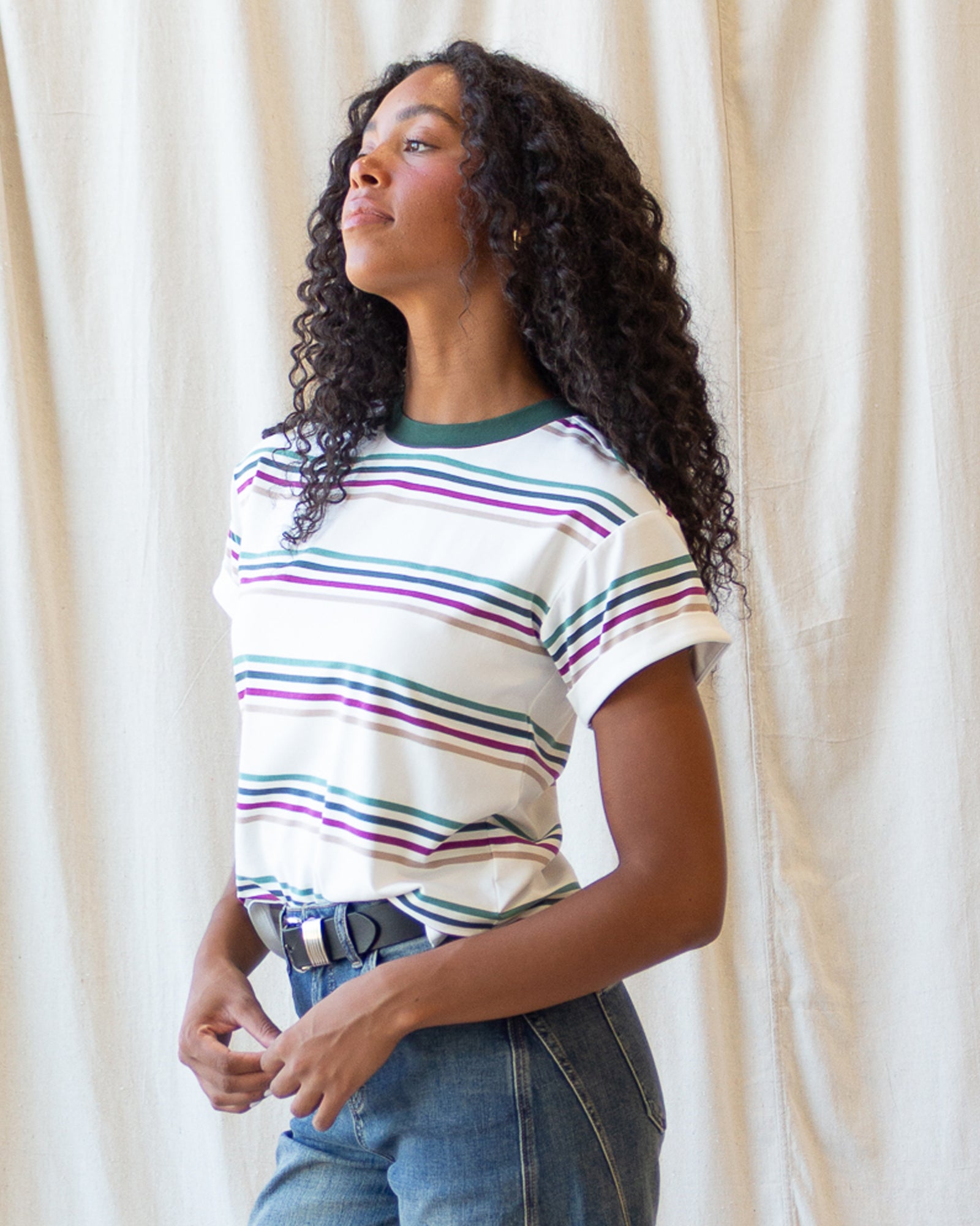 A woman with long curly black hair models a white short-sleeve t-shirt with thin horizontal stripes in green, purple, and blue, paired with blue jeans and a black belt, standing against a draped cream fabric backdrop.
