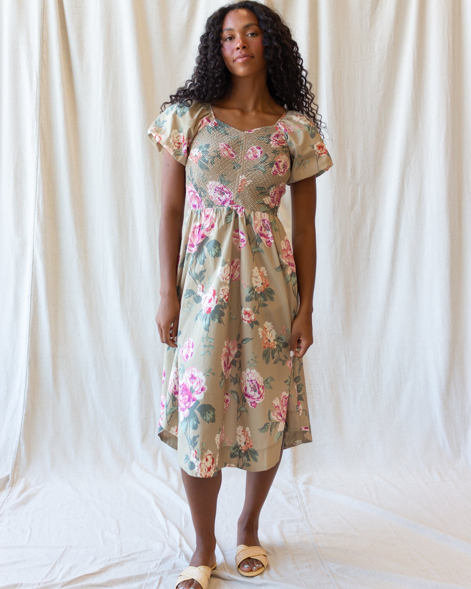 A woman modeling a short-sleeve midi dress with a beige base and a pink, green, and blue floral print, standing against a draped white fabric backdrop.