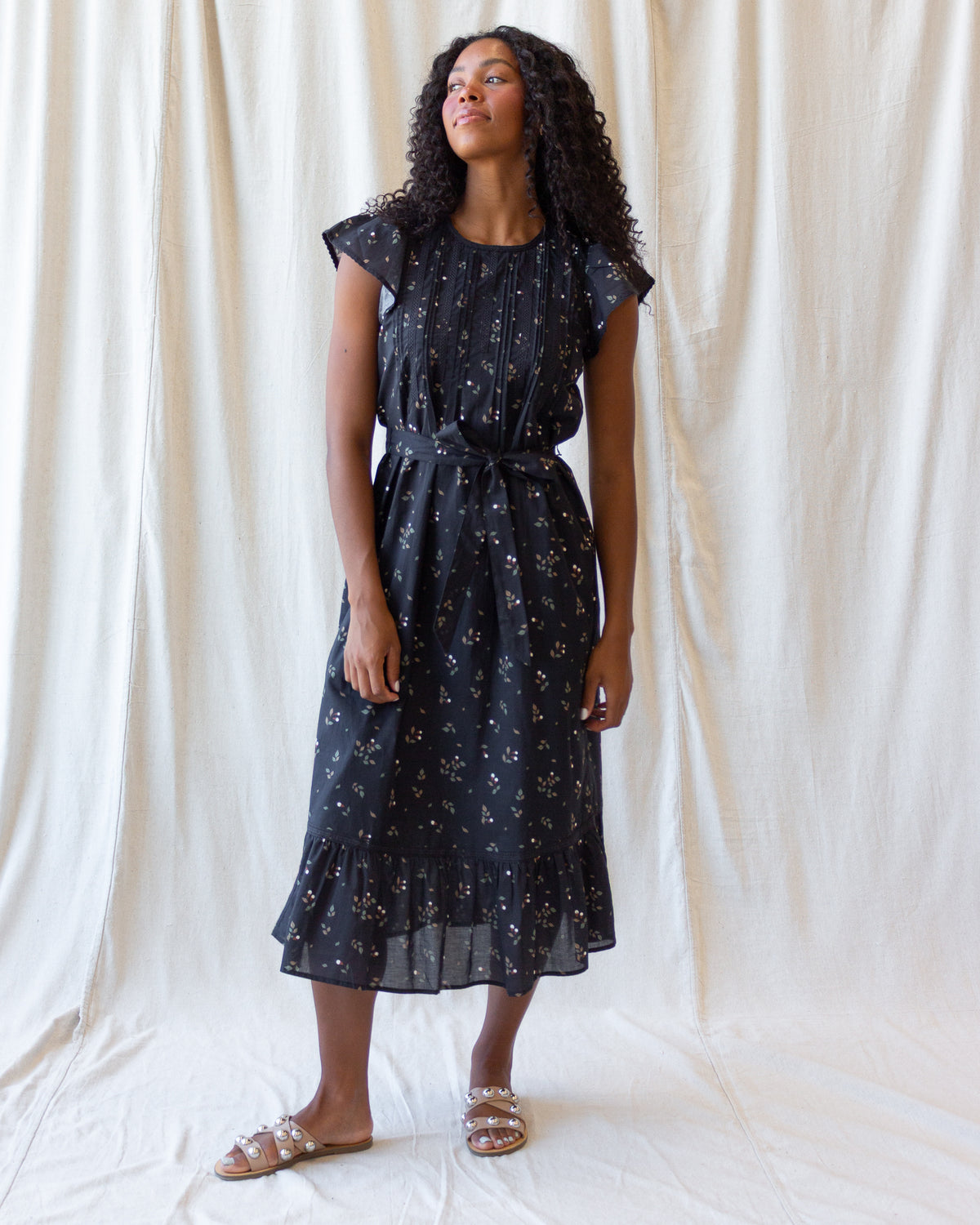 A woman modeling a dark navy midi dress with a delicate white and gold floral print, standing against a draped off-white fabric backdrop.