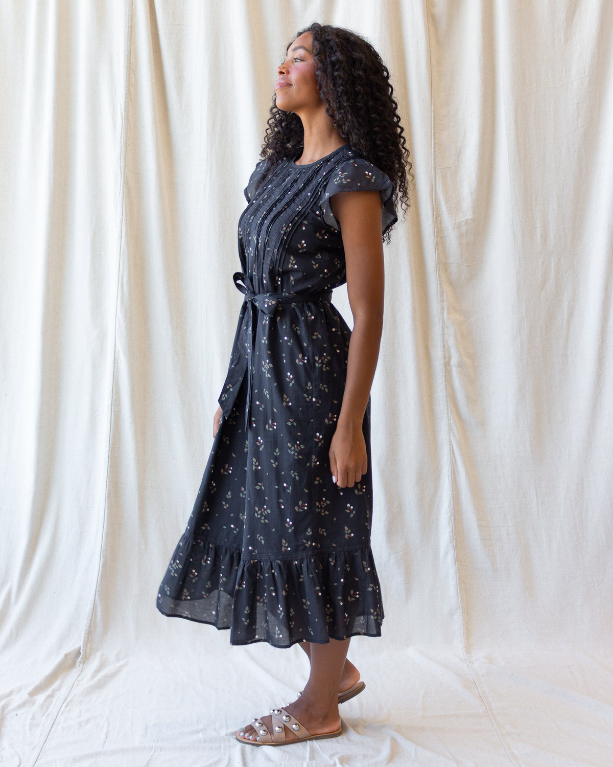 A woman wearing a dark navy midi dress with a white floral print and a matching waist tie, standing in front of a draped cream fabric backdrop.