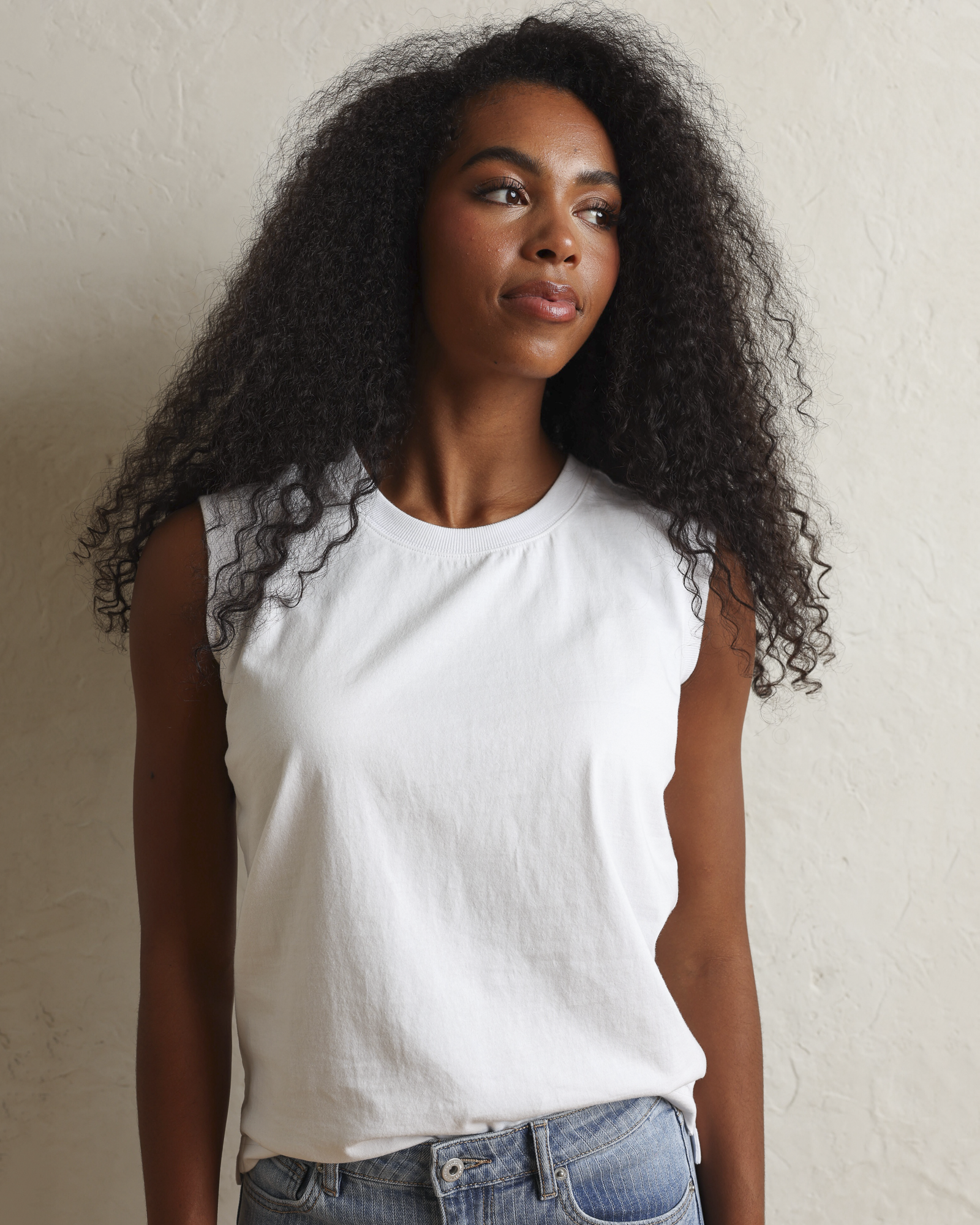 A woman with long, curly black hair wearing a white sleeveless top and light blue jeans, standing against a textured off-white wall.