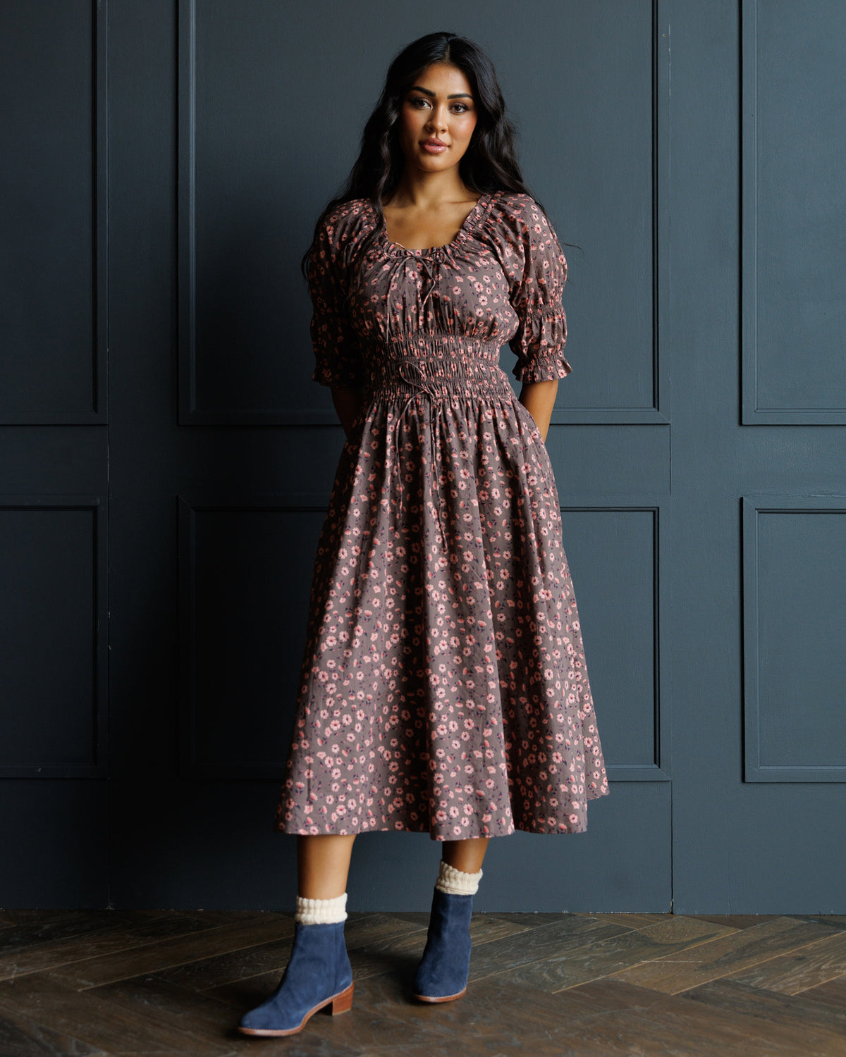 A woman stands against a dark paneled wall wearing a dusty rose midi dress with a pink floral pattern, puff sleeves, and a smocked waist. She is wearing blue suede ankle boots with white socks.