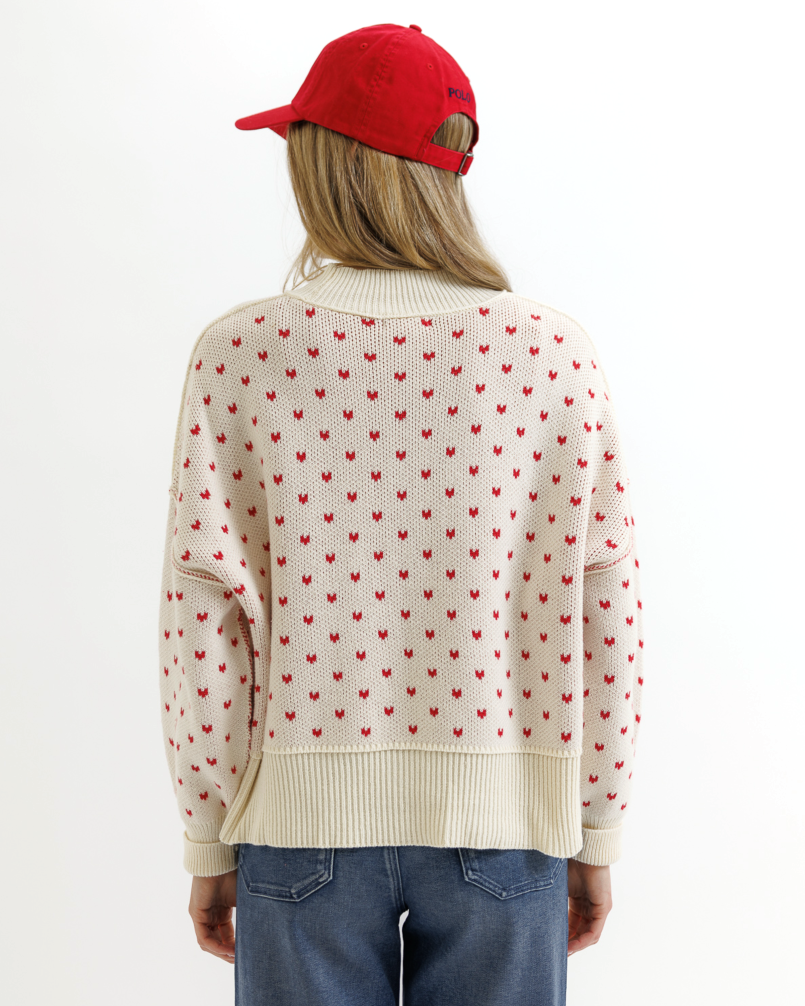 Woman wearing a cream sweater with red heart patterns, blue jeans, and a red cap on a white background