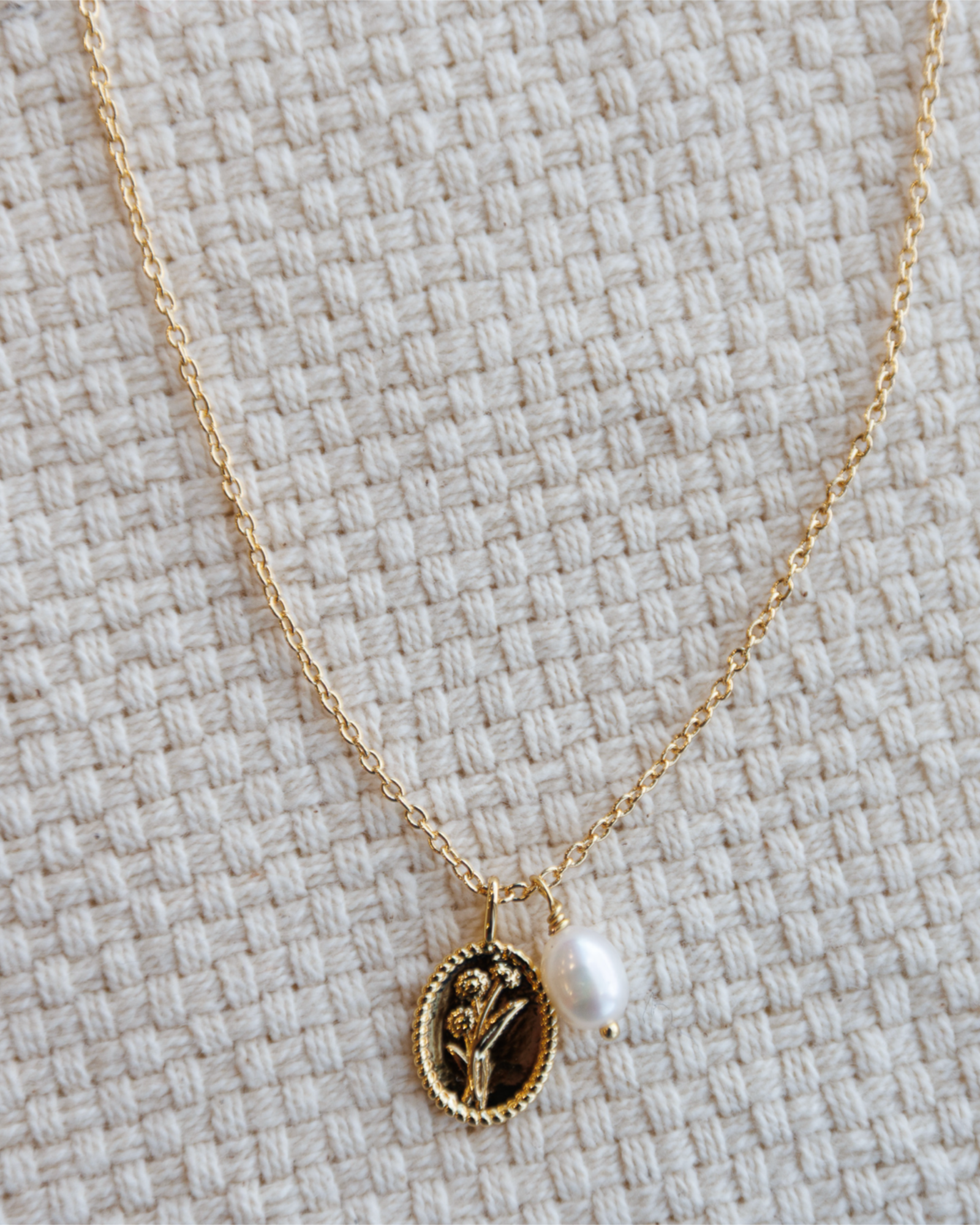 Gold necklace with a flower pendant and pearl charm on a textured beige surface