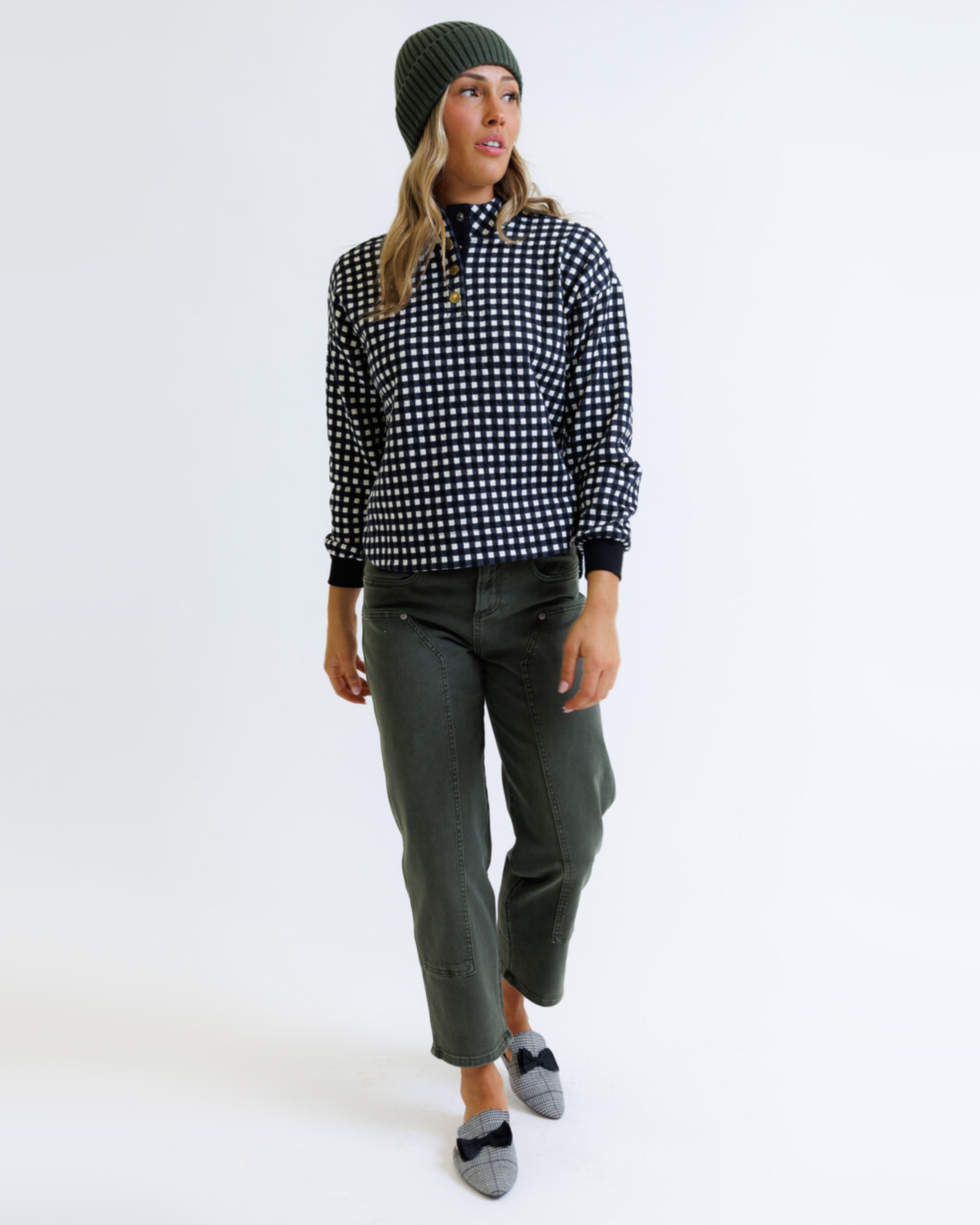 Woman wearing a black and white checkered pullover, green beanie, and green pants on a white background