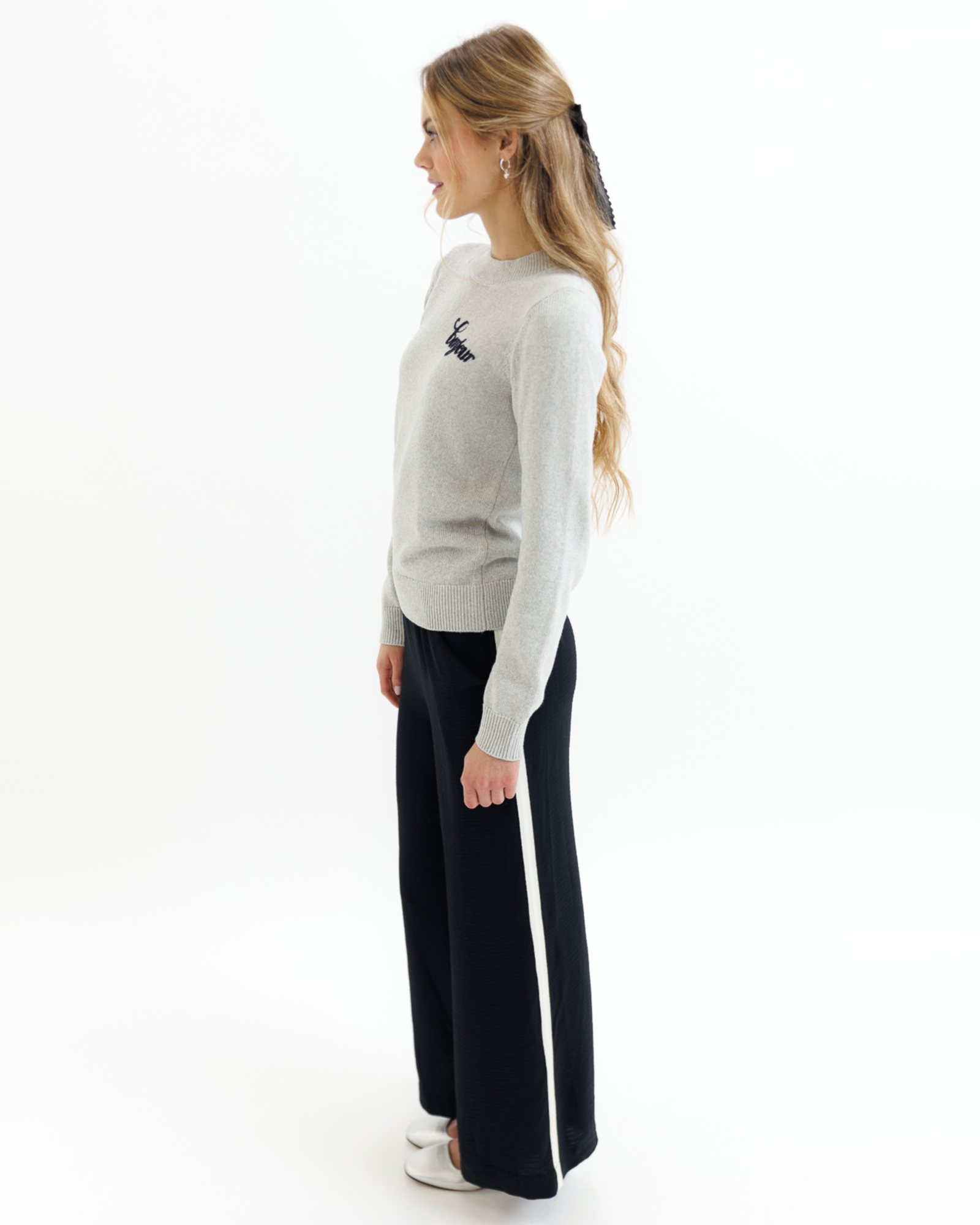 Woman wearing a gray sweater with black embroidered text that says 'bonjour' and black pants on a white background