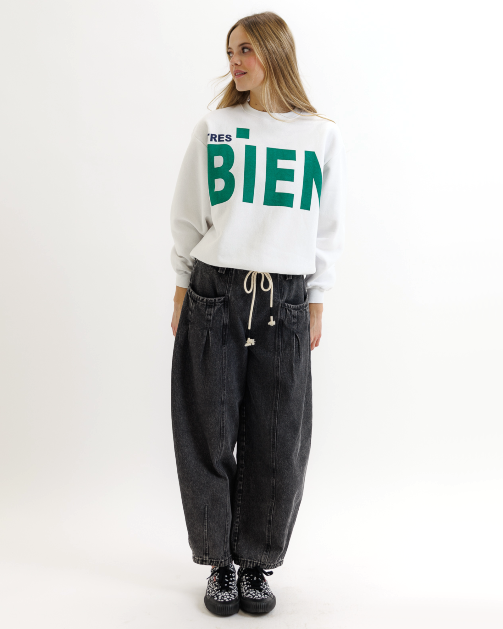 Woman wearing a white sweatshirt with green text that says 'TRES BIEN' and black pants on a white background
