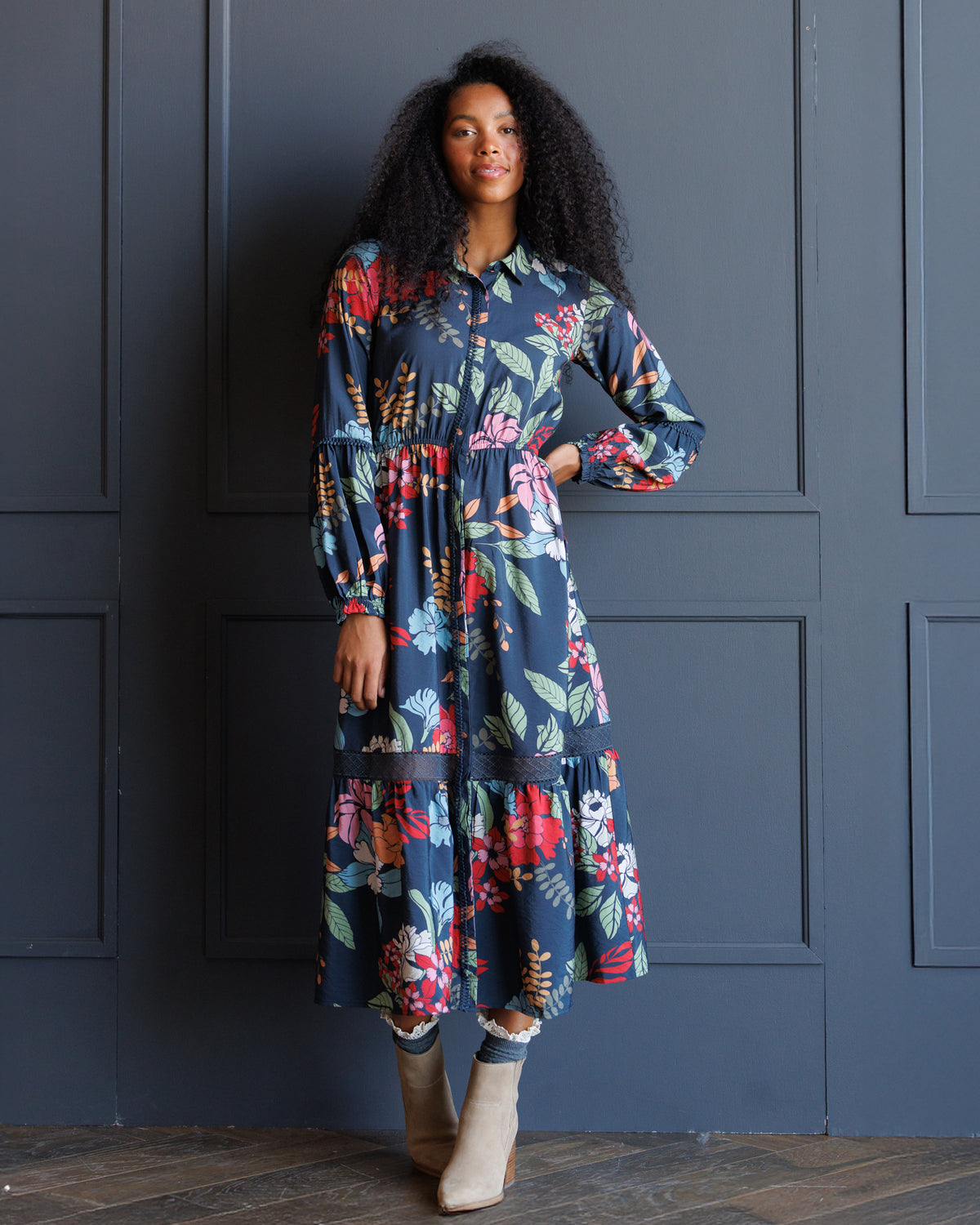 A woman with curly black hair models a long-sleeved navy blue midi dress with a colorful floral print, standing against a dark blue paneled wall.
