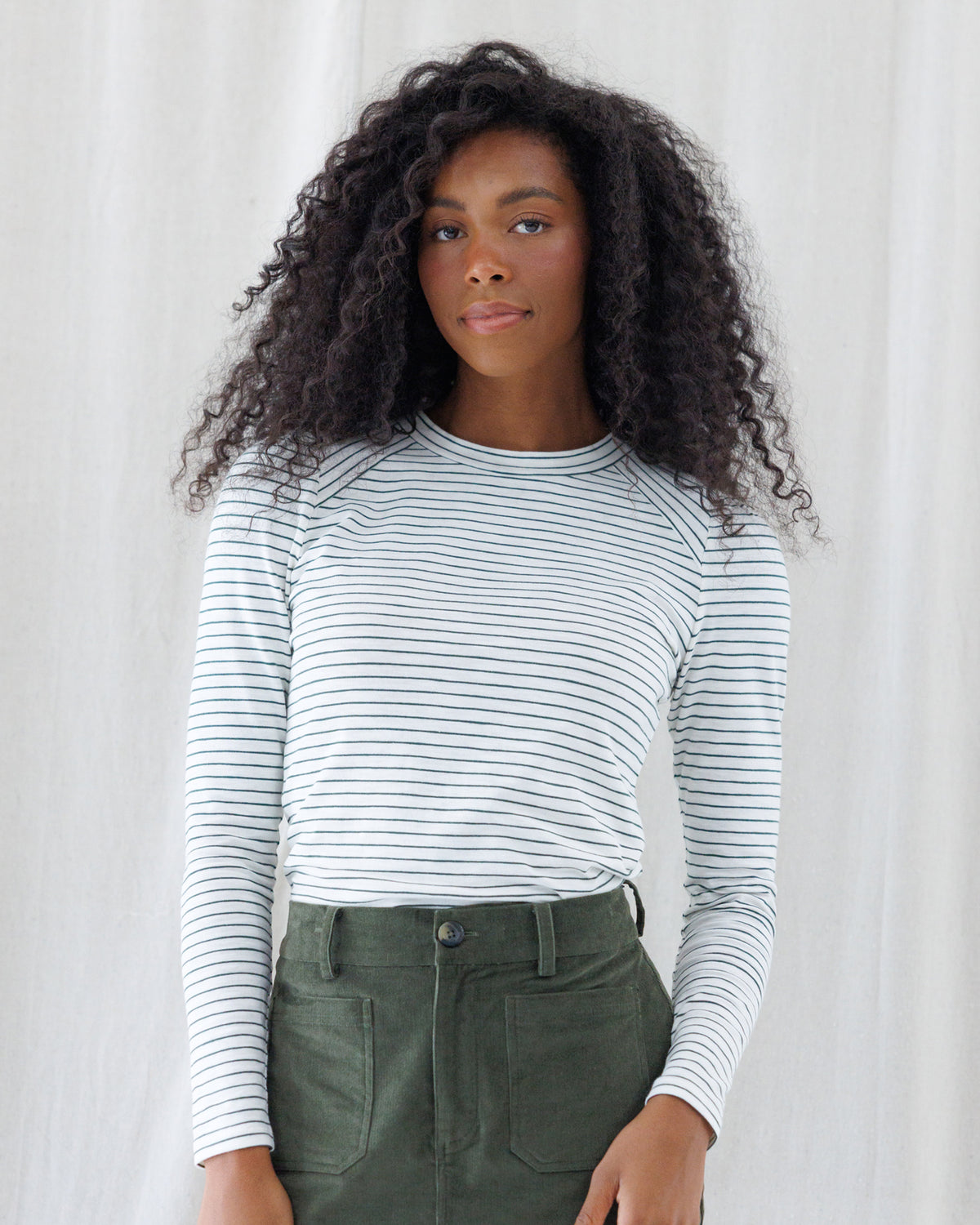 A woman with curly black hair wearing a white long-sleeve raglan top with thin dark horizontal stripes and olive green pants, standing against a white curtain background.