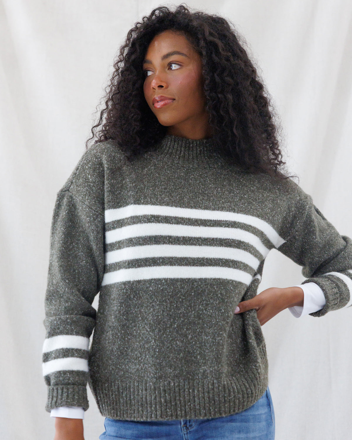 A woman wearing a heathered olive green knit sweater with three horizontal white stripes across the chest and white striped cuffs, paired with blue jeans, set against a white draped fabric background.
