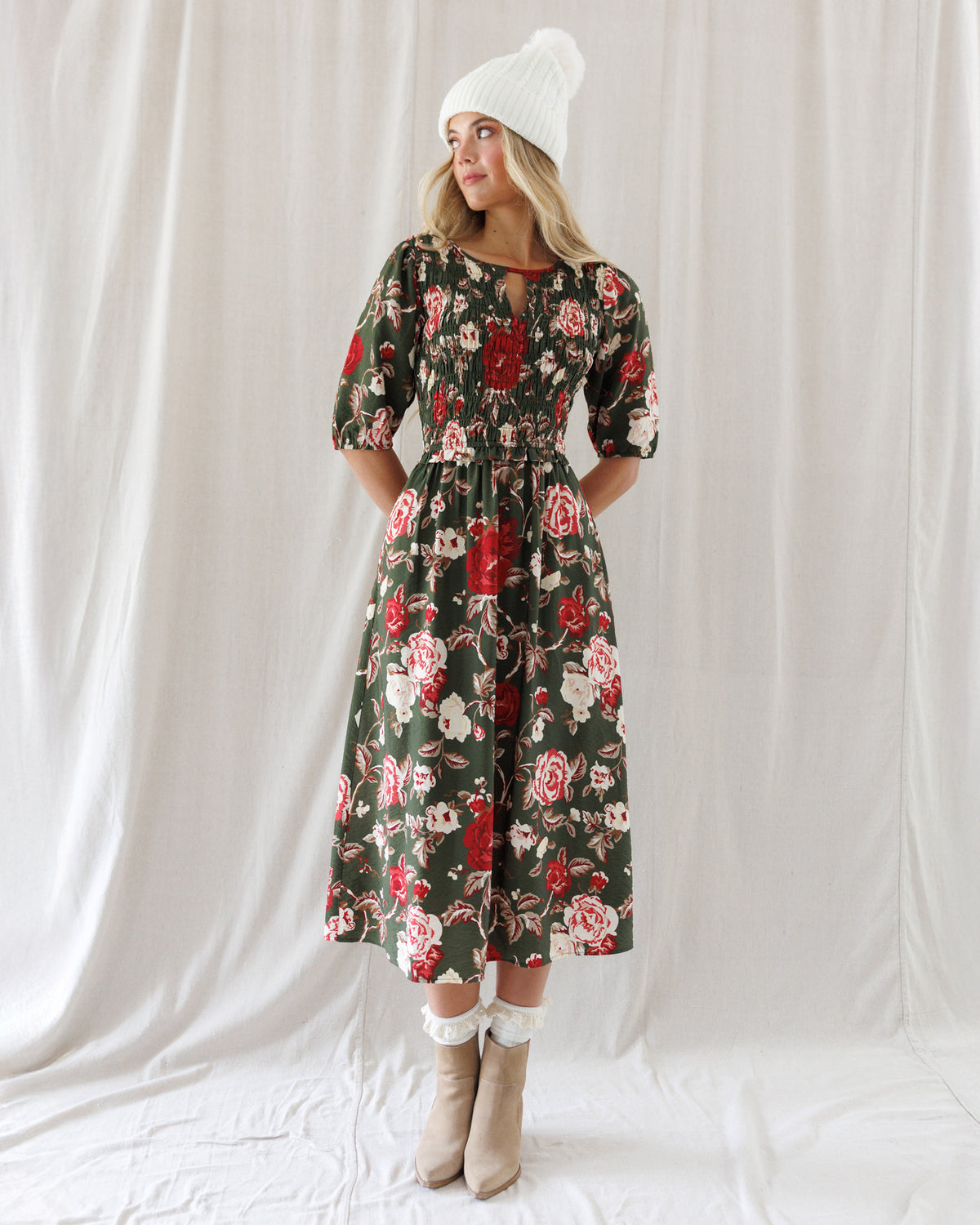 A woman stands against a white draped fabric backdrop wearing a dark floral midi dress with puff sleeves, a white knit beanie, white socks, and beige ankle boots.