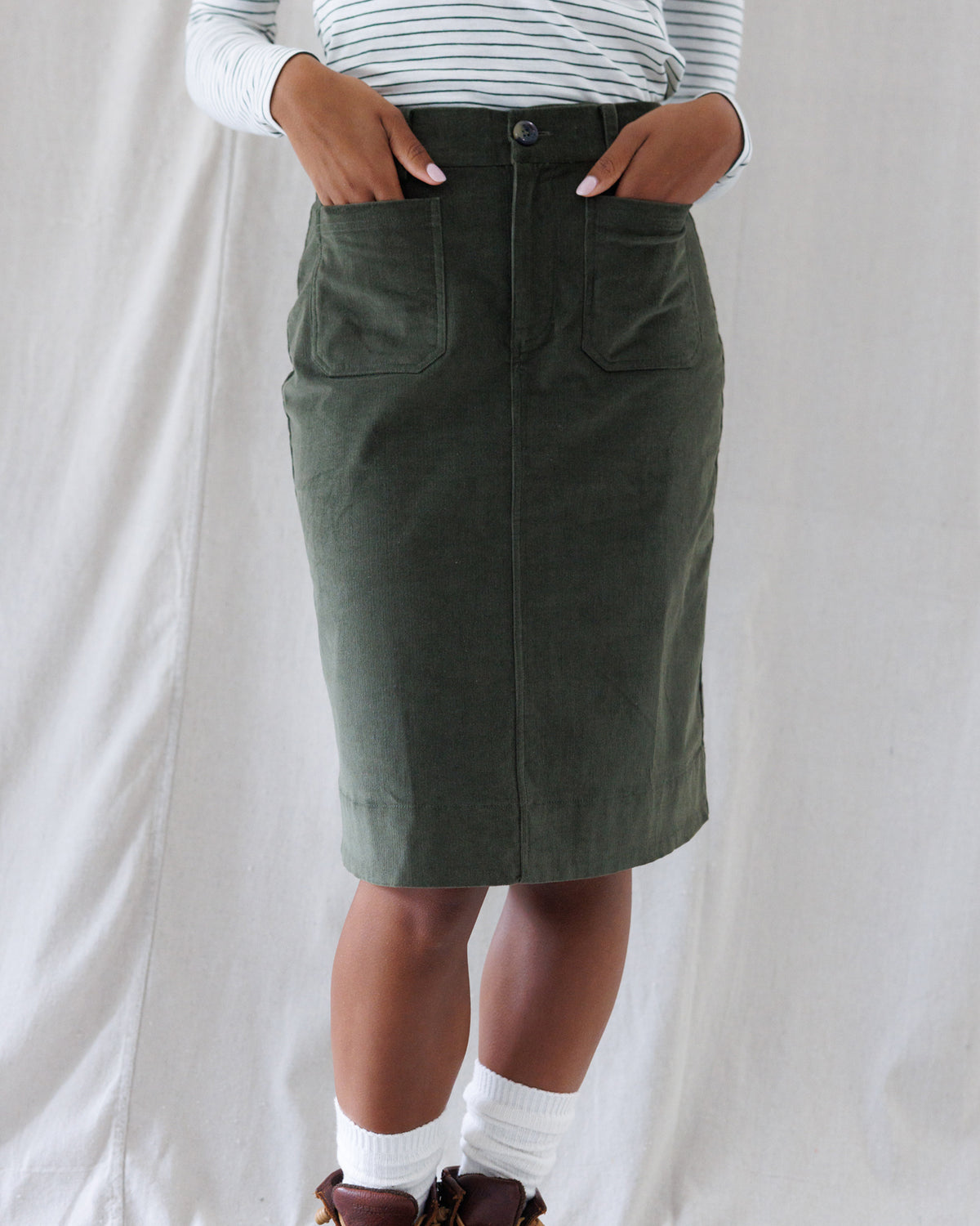 An olive green corduroy pencil skirt with front patch pockets, worn by a model with a striped long-sleeve shirt and white socks against a white fabric backdrop.