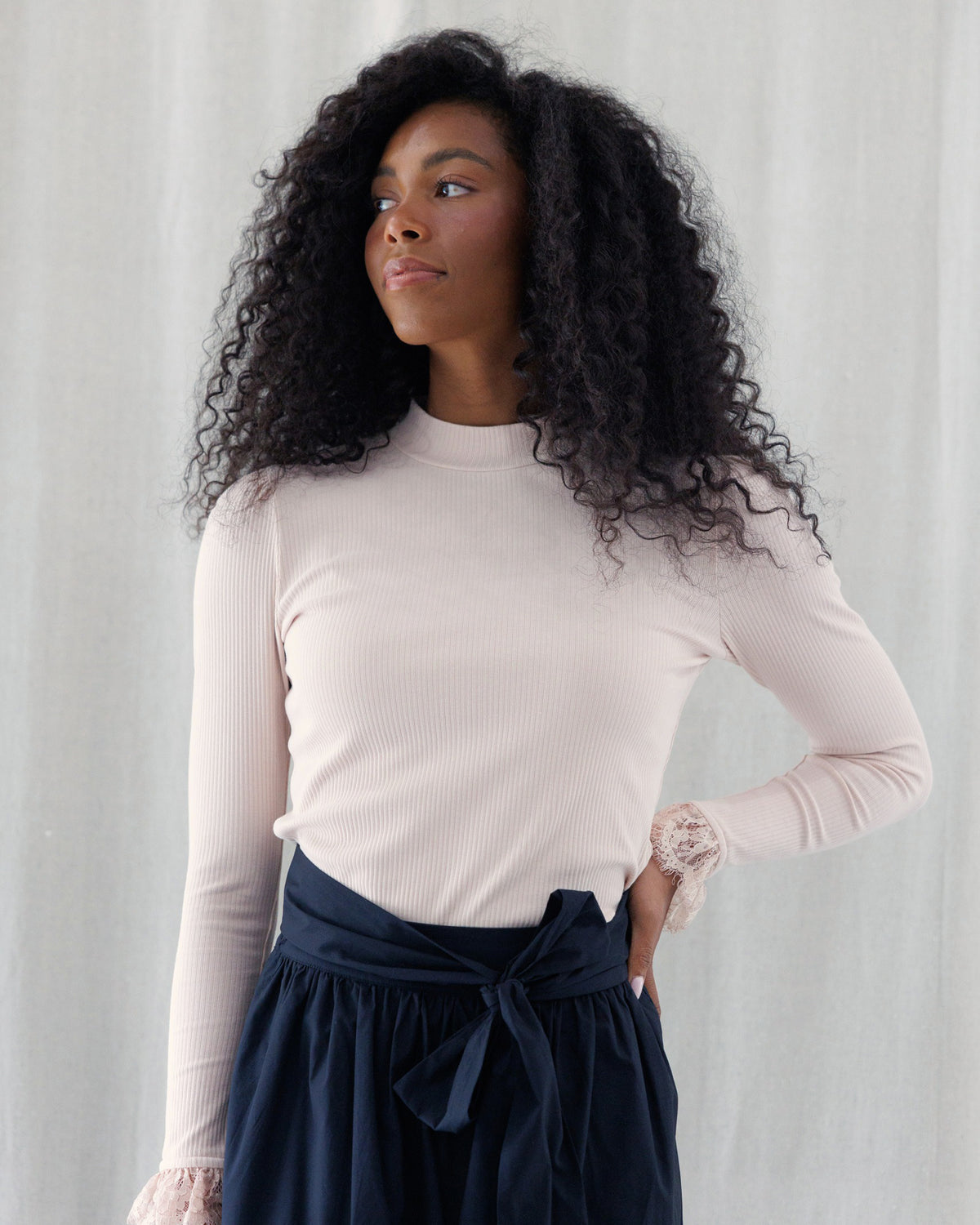 A woman with long, curly black hair wearing a light pink ribbed long-sleeve mock neck top and a navy blue skirt with a tie waist, standing against a light gray curtain background.