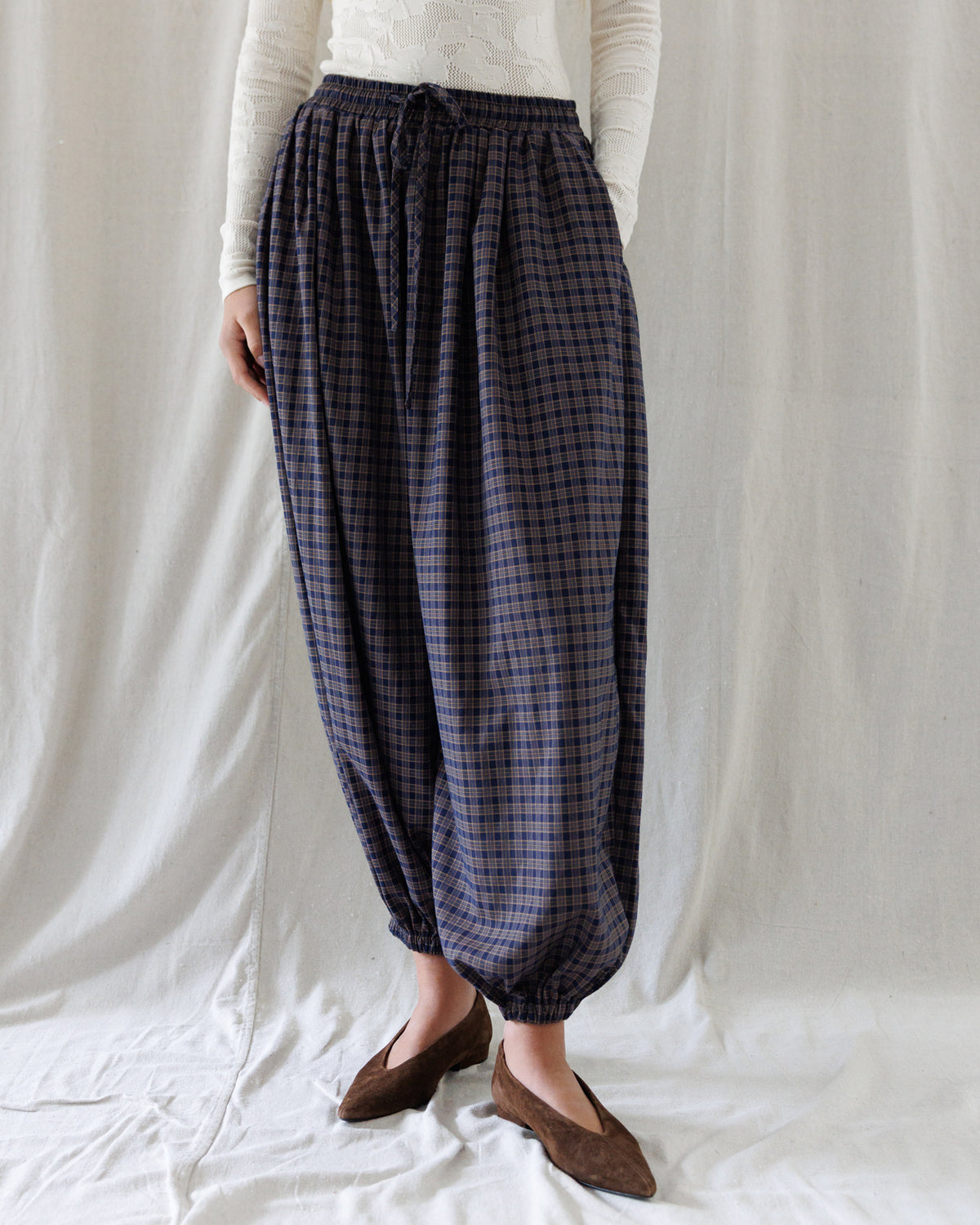 A person wearing loose, high-waisted harem-style pants with a dark blue and black checkered pattern, paired with a white long-sleeve top and brown slip-on shoes, standing against a white fabric backdrop.